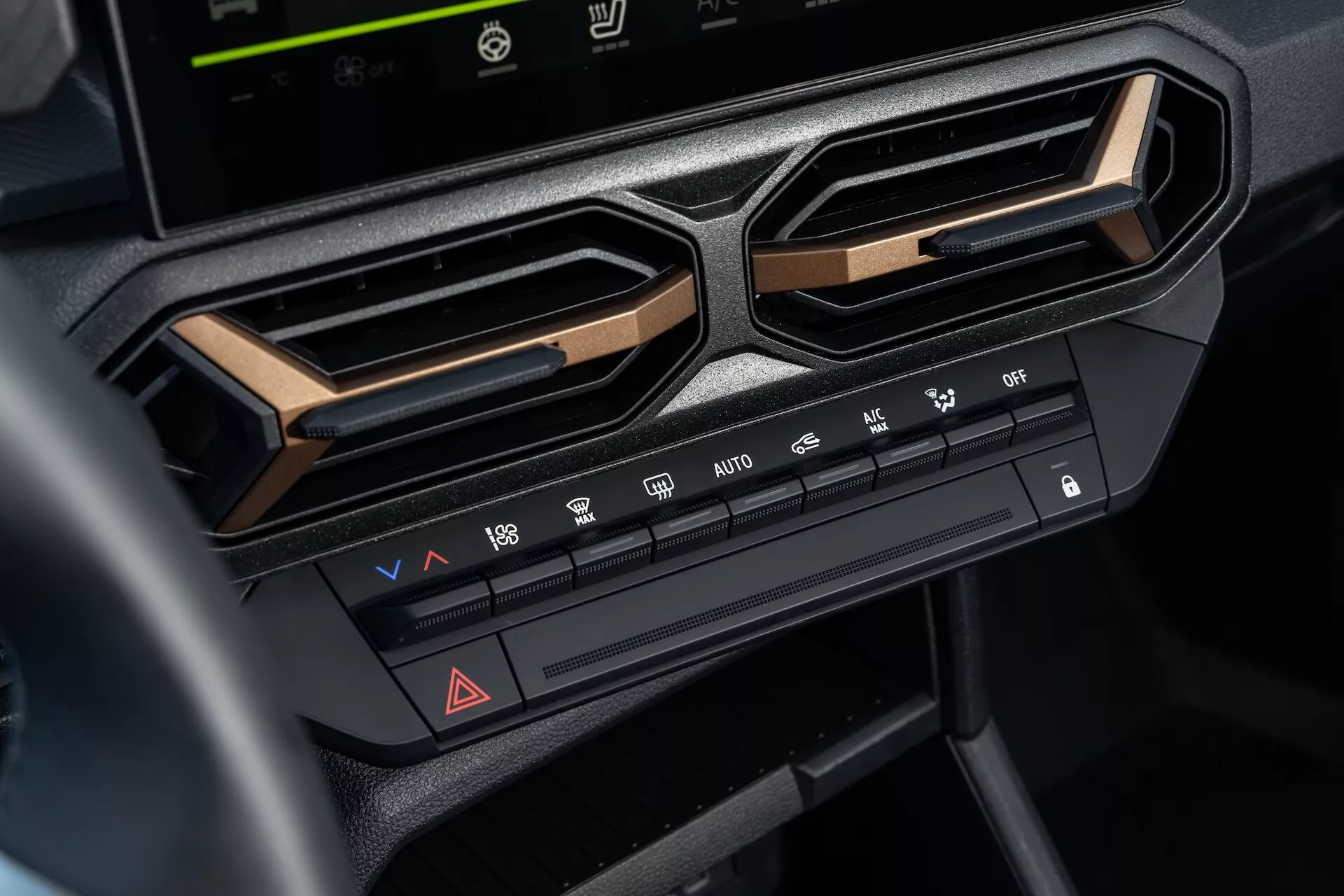 Närbild av klimatkontrollsystemet på 2024 Dacia Duster 3:e generationens centrala instrumentpanel under underhållningsskärmen, som framhäver justeringar av temperatur och fläkthastighet, luftfördelningsfunktioner, avfrostningsfunktioner, varningsljusknap, och luftkonditioneringsalternativ, inramade av vinklade luftventiler och accentuerade med metallisk dekor för en modern interiör estetik
