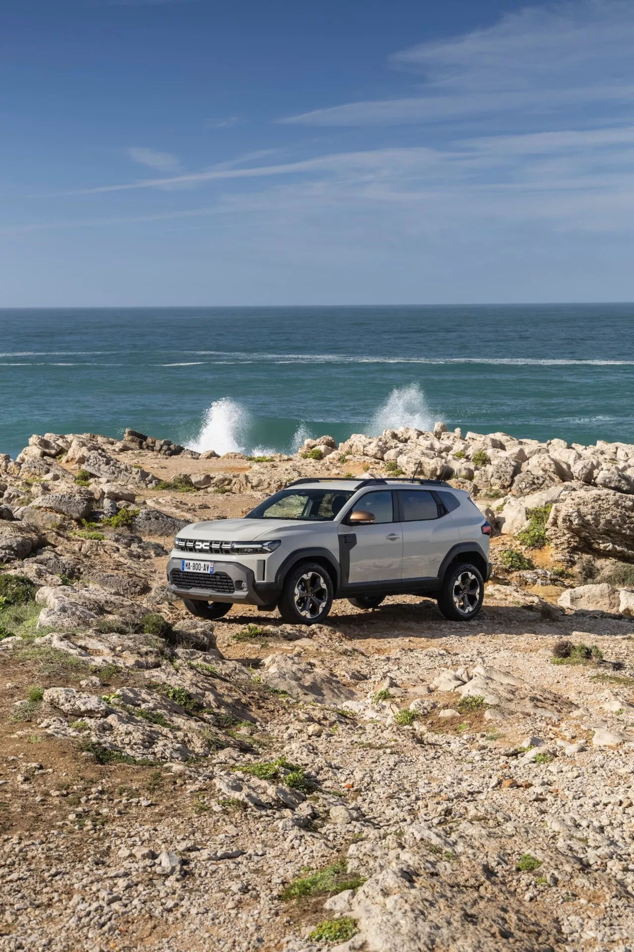 Sport utility-fordon Dacia Duster 2024 (3:e generationen) parkerat på klippig terräng med kustbakgrund, med kontrasterande färgdesign, stora lättmetallhjul, LED-belysning, hög markfrigång och takräcken, fångat i dagsljus under klar himmel