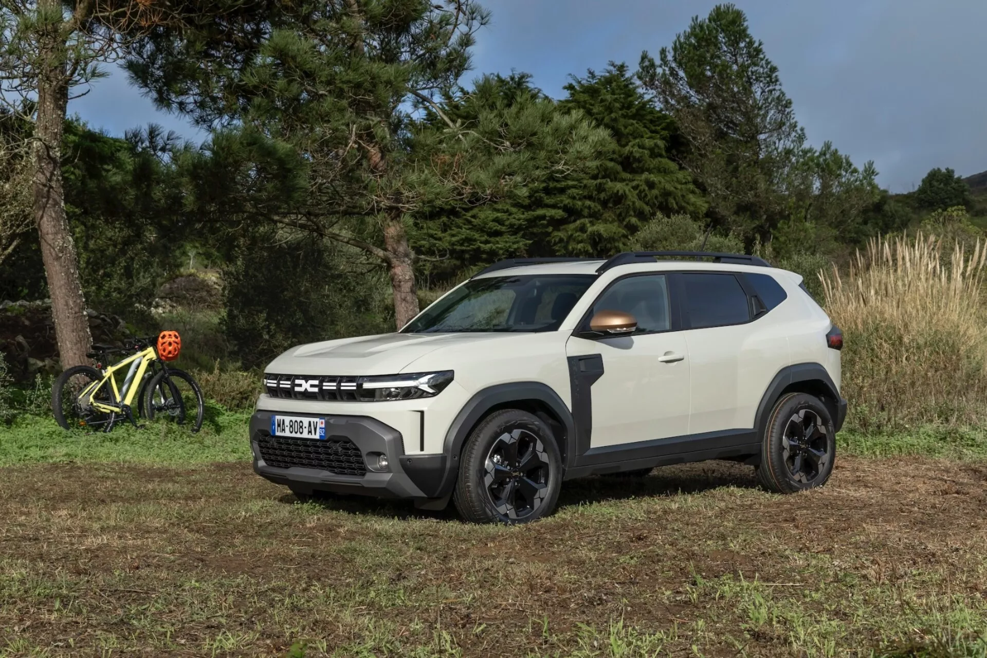2024 Dacia Duster 3:e generationen i vitt med svarta detaljer, fångad från ett tre fjärdedels frontperspektiv som visar dess robusta design och aerodynamiska profil i en naturlig, möjligtvis off-road miljö med molnigt himmel