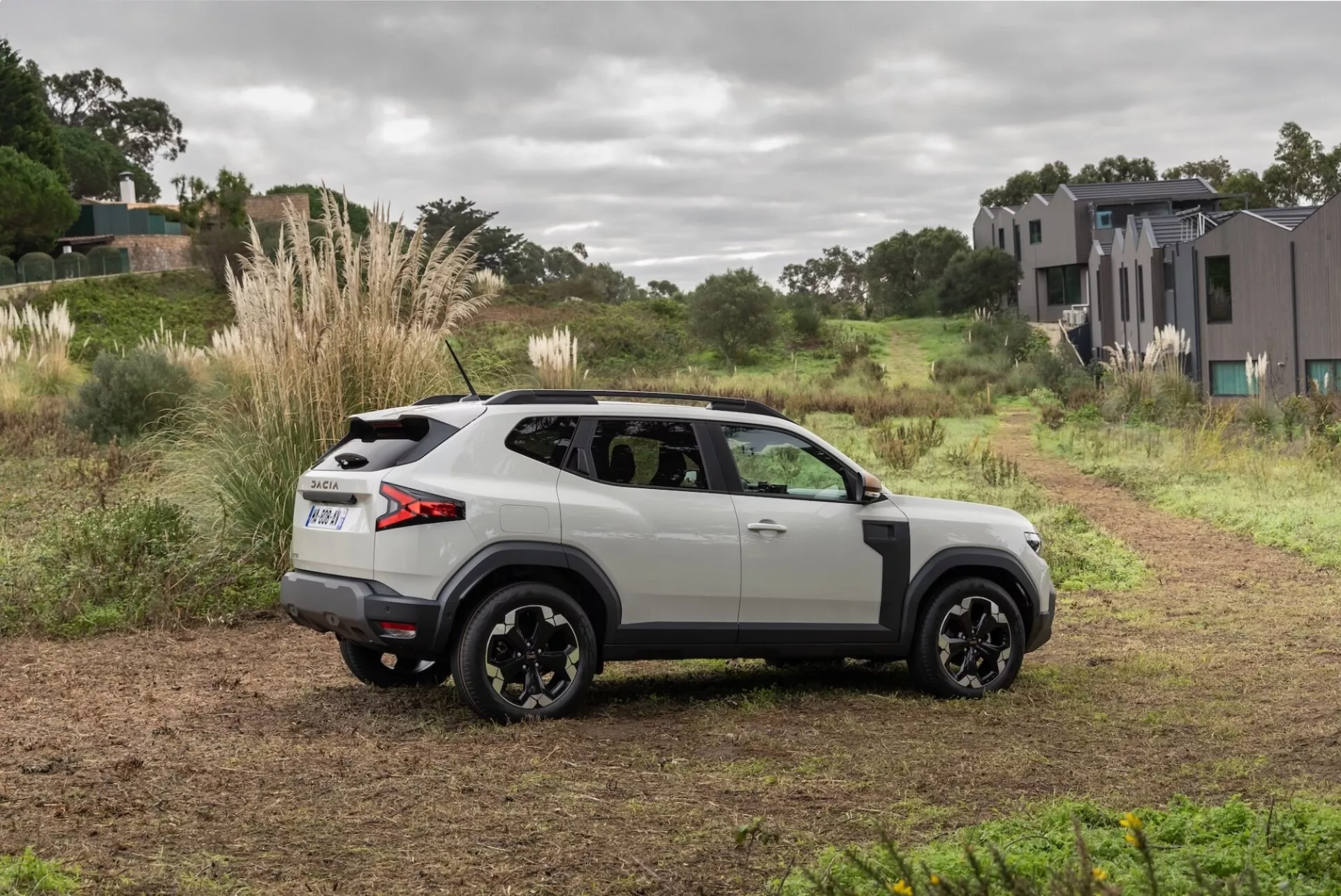 Dacia Duster 2024, 3:e generationen, vit med svarta detaljer, parkerad på en lantlig grusväg med en vy från sidan bakifrån som framhäver dess aerodynamiska profil, moderna LED-bakljus, och svarta lättmetallfälgar, designad för både stad och terrängäventyr, under mjukt naturligt ljus