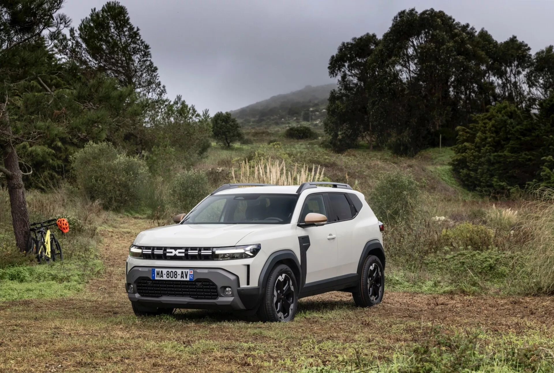 Dacia Duster 3:e generationen 2024 positionerad utomhus på en terräng lämplig för terrängkörning med molnigt belysning, som visar sin främre grill, LED-strålkastare, tvåfärgat färgschema och moderna SUV-designfunktioner.