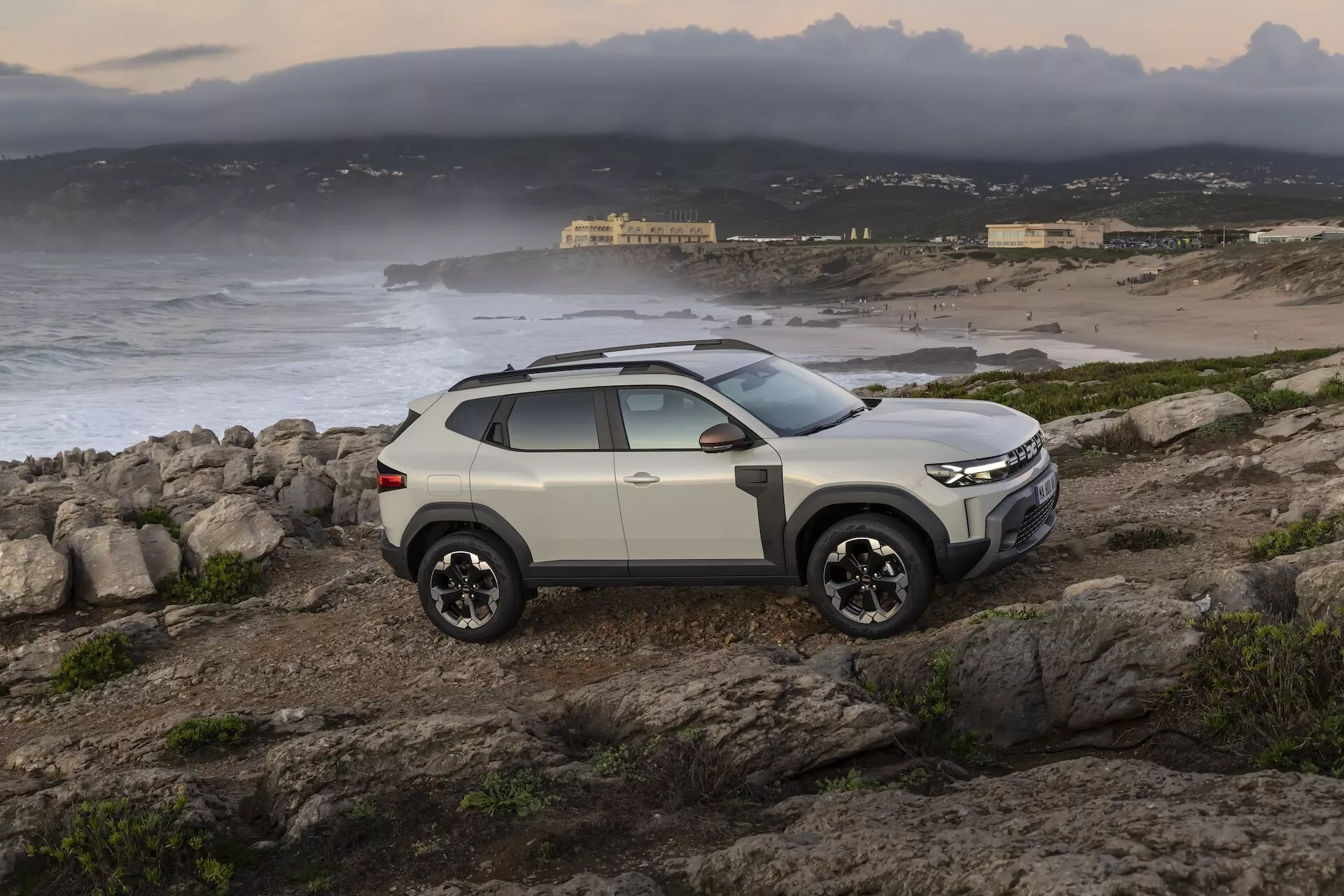 Modernt SUV Dacia Duster 3:e generation 2024 parkerat på ojämn, klippig terräng med kustbakgrund, visar frontgrillen, strålkastarna, stötfångaren, motorhuven och förarsidans profil med terrängdäck, från en tre fjärdedels främre vinkel