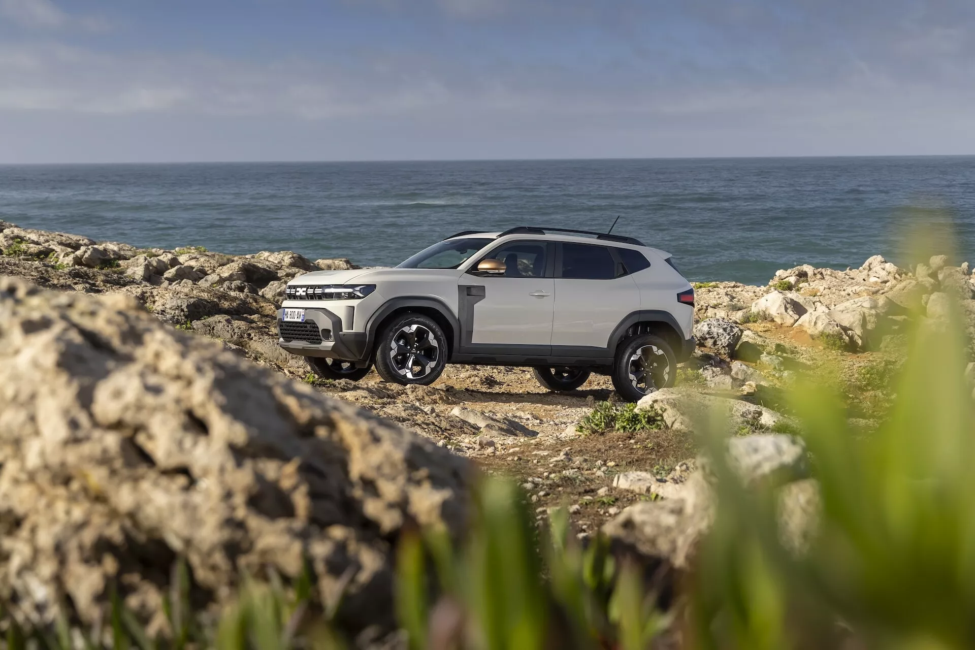 Dacia Duster 3:e generationen från 2024 parkerad på en klippig kust med havet i bakgrunden, som visar terrängkörningsförmåga och en modern, robust design med tvåfärgad lack och svarta lättmetallhjul