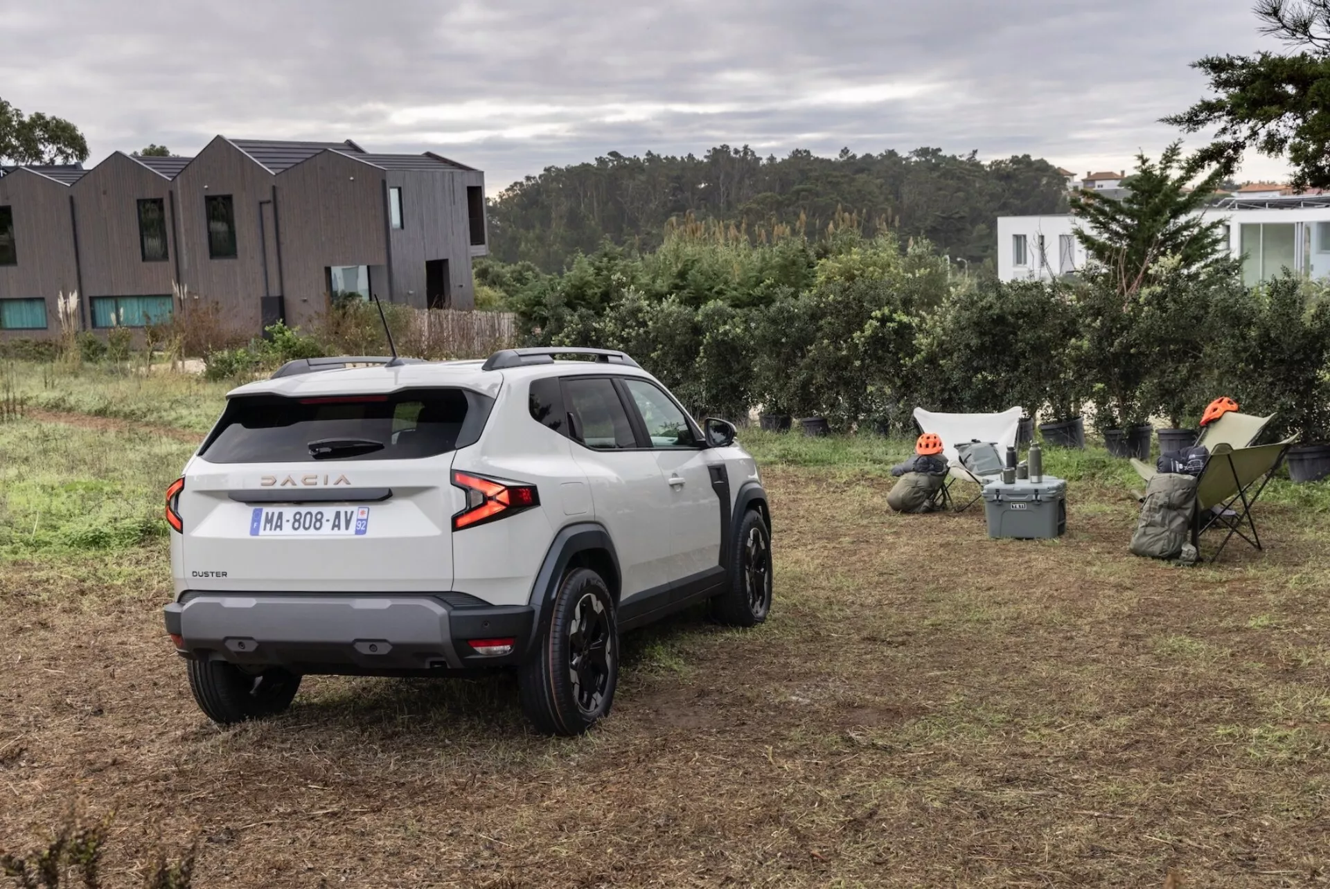 Trebjärtad bakvy av en silver Dacia Duster SUV 2024 parkerad på en gräsmatta, framhävande bakljus, bakfönster, takspoiler och legeringshjul, vilket antyder lämplighet för lätt terrängkörning, med europeisk registrering och ett lugnt landsbygdsbakgrund.
