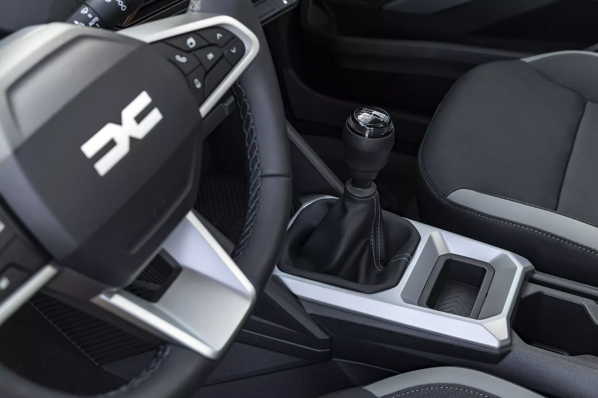 Närbild av interiören på förarsidan av Dacia Duster generation 3 2024, med en multifunktionell ratt med silverdetaljer och kontrollknappar, en manuell växelspak och sportiga läderimitationssäten.