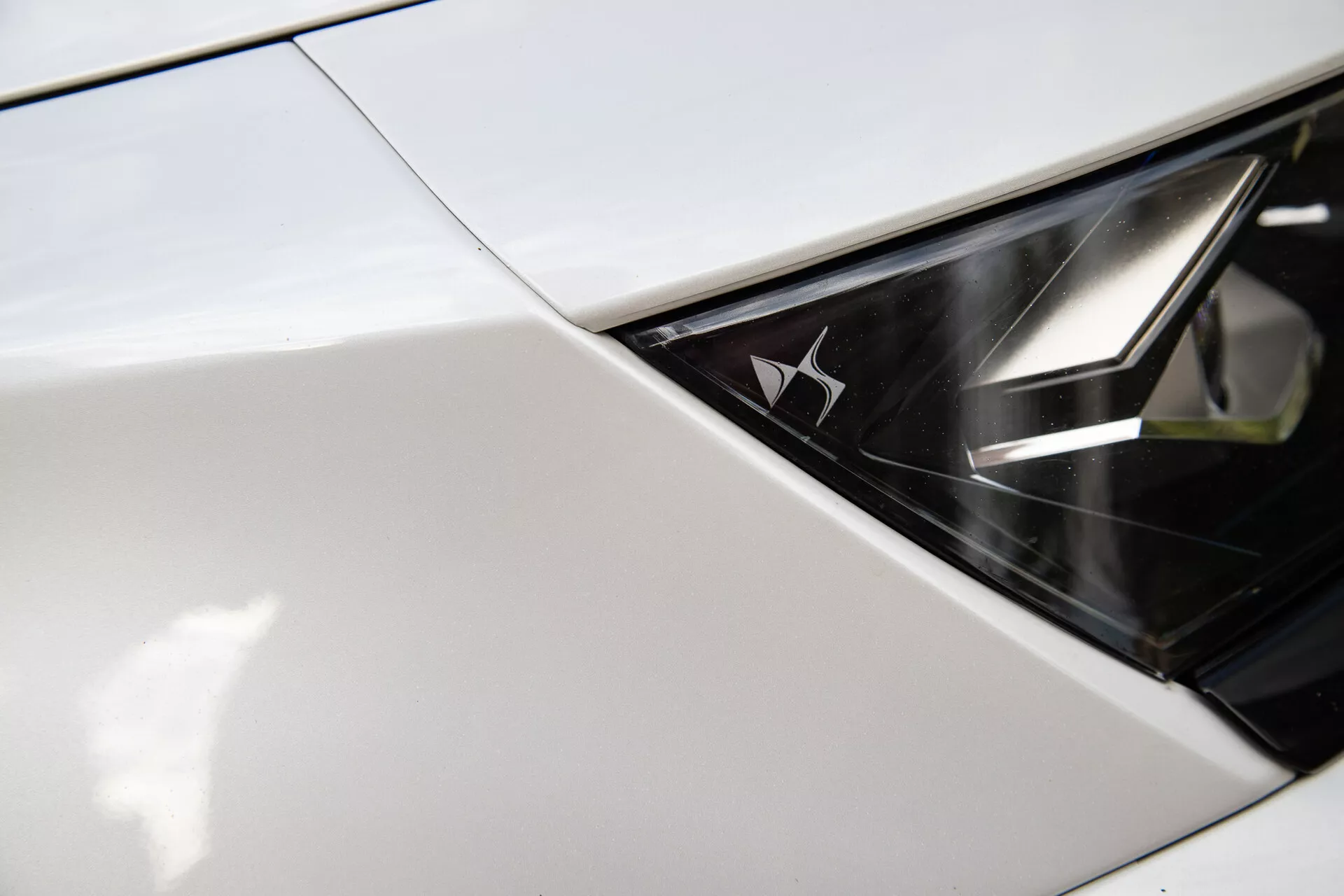 Närbild på DS Automobiles DS-7 2022:s strålkastare som visar skärningspunkten mellan skärm eller huva med strålkastarmonteringen, visar upp en glänsande, monokrom design och ett varumärkesemblem inuti strålkastaren, antyder modern lyx och avancerad LED-belysningsteknologi.