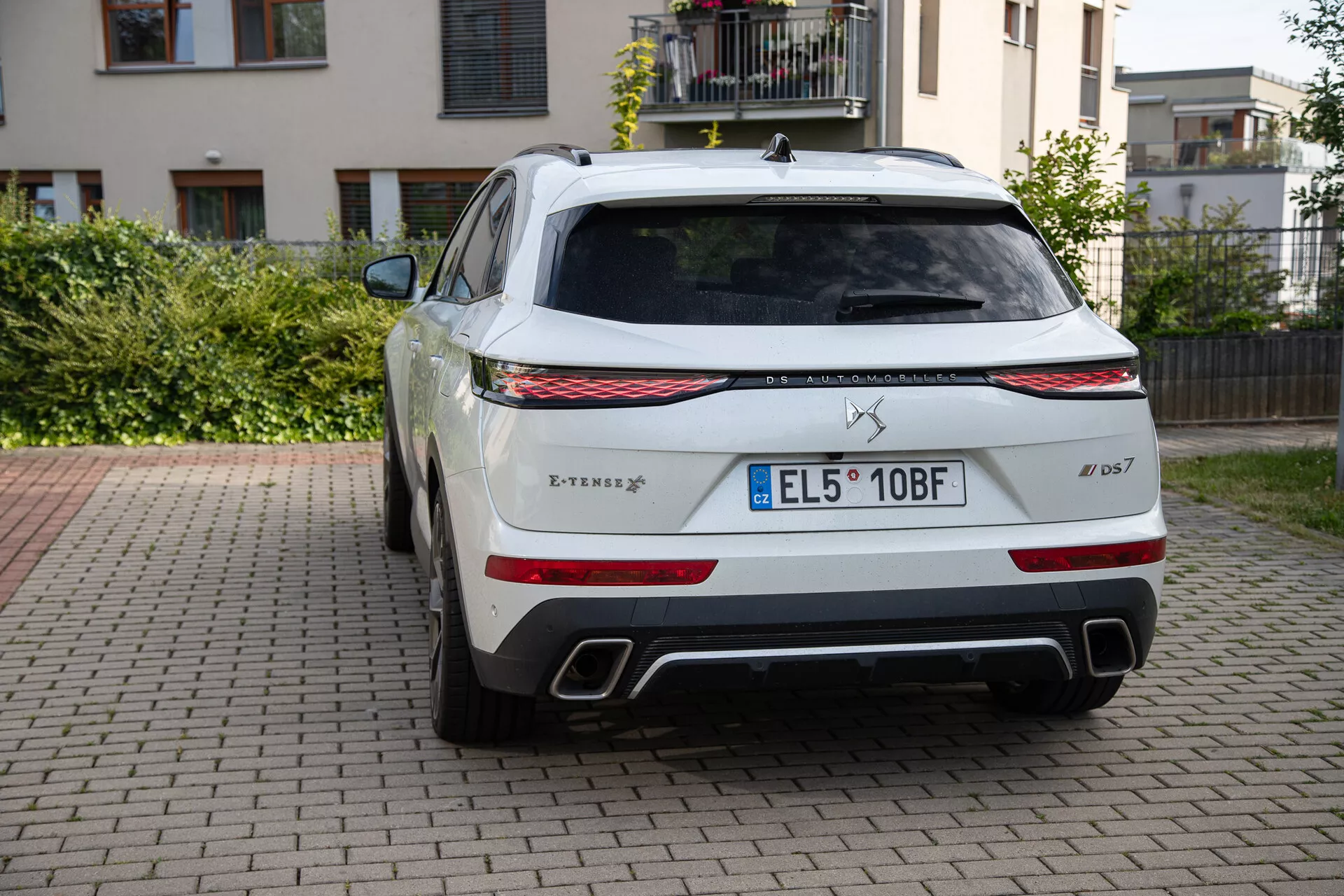 Bakvy av en DS Automobiles DS 7 2022 parkerad på asfalt med bostadsbyggnad och grönska i bakgrunden, med trident logotyp, Levante modellnamn, S trim märke, LED bakljus, dubbla kromade avgasrör, svart diffusor, stort bakfönster med torkare, europeisk registreringsskylt, parkeringssensorer, Q4 fyrhjulsdrift märke, och hajfena antenn.