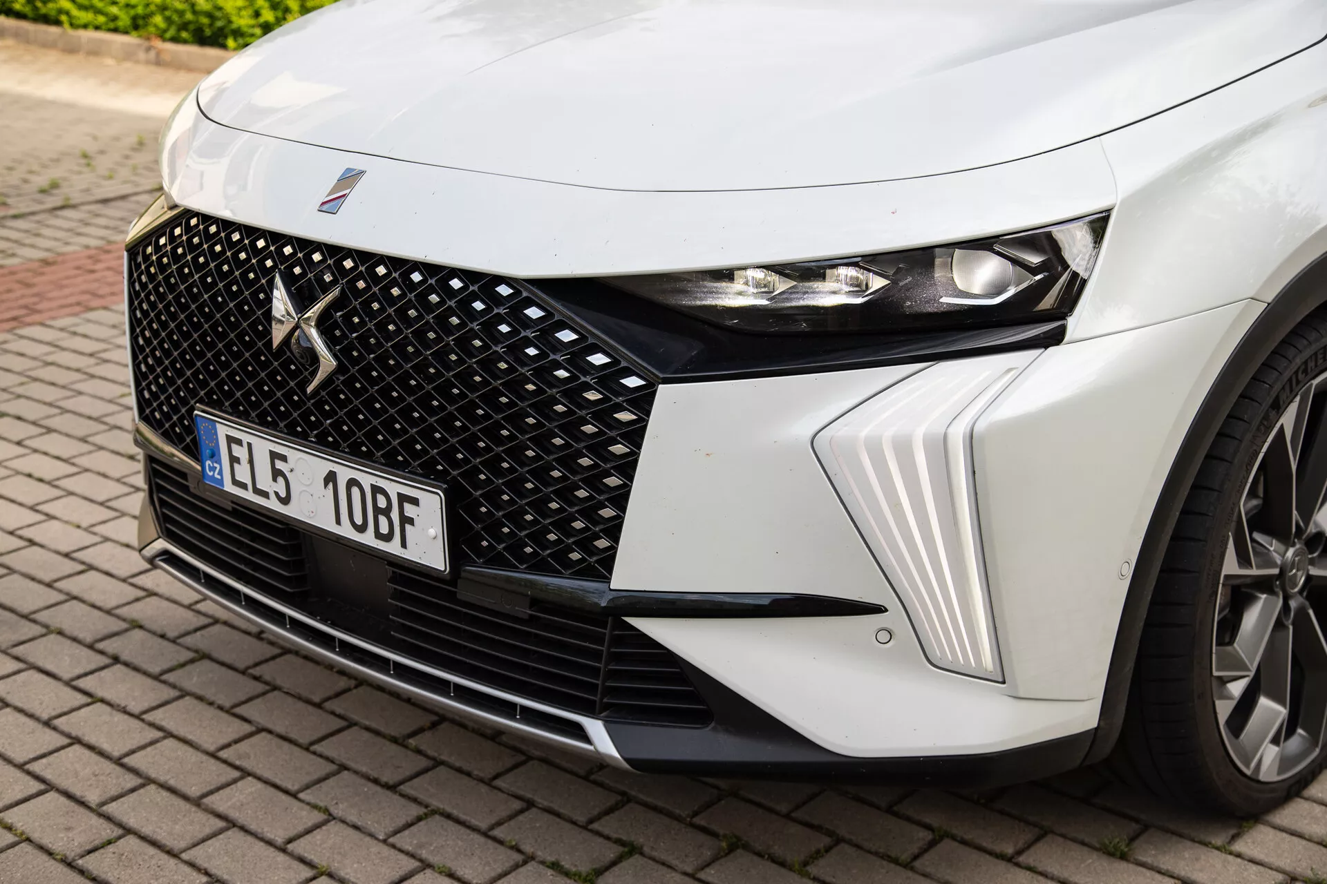 Närbild av det främre vänstra kvadranten på DS Automobiles DS-7 2022 med detaljerat fokus på LED-strålkastaren, hexagonalt grilldesign med centralt märkesmärke och dynamiska huvgångar tillsammans med en glimt av legeringshjulet