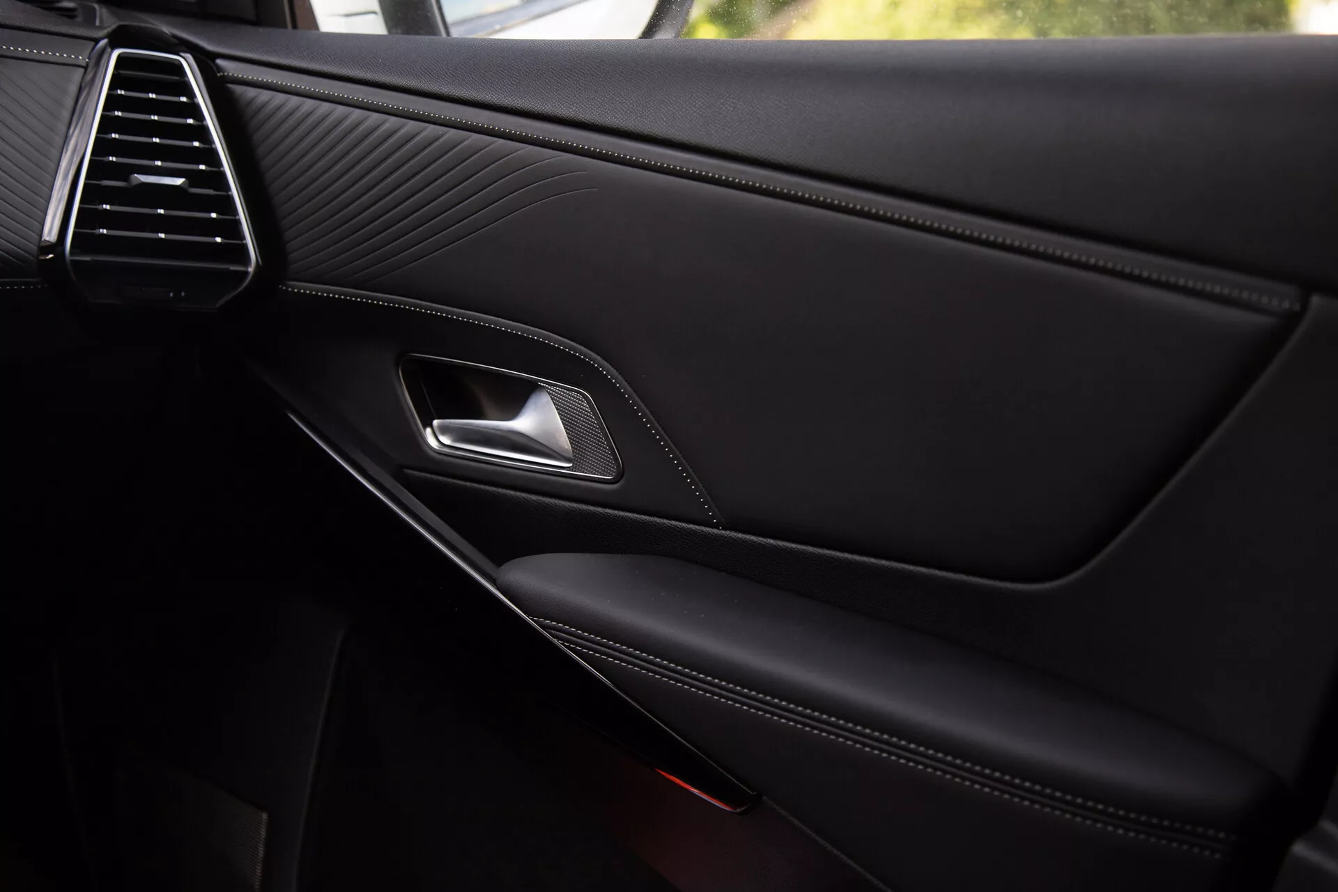 Närbild av DS Automobiles DS-7 2022 interiördörrpanel som visar den eleganta luftventilen, stygnad lädertextur, kromad dörrhandtag, ergonomisk låsvred, kontraststygnad armstöd och integrerade förvaringsfacket med delvis synlig högtalargaller, vilket återspeglar premiumkvalitet och design detaljer.