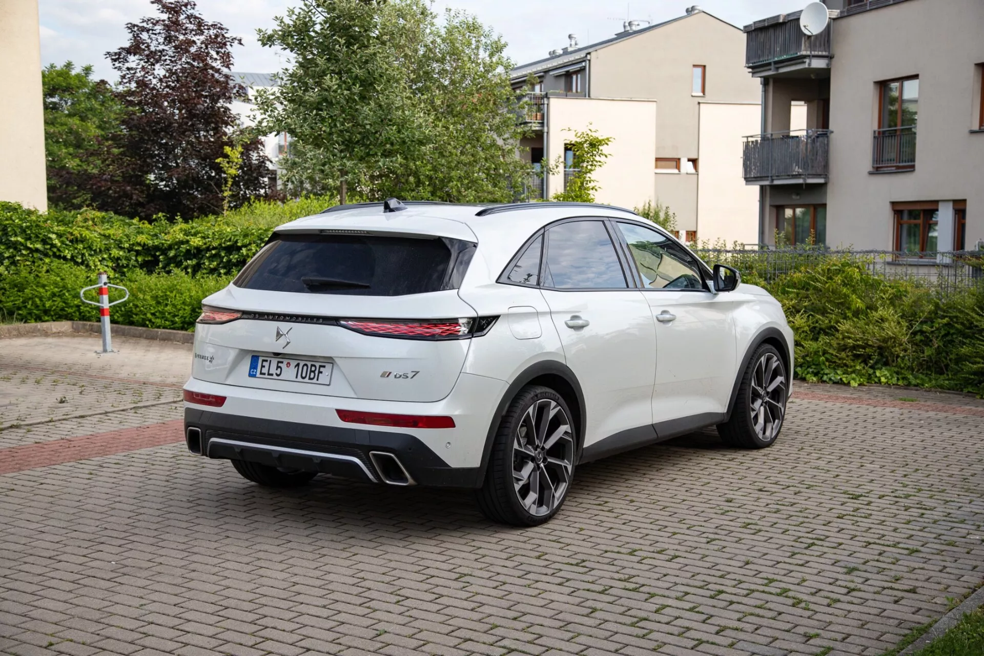 Vit SUV DS Automobiles DS-7 från 2022 parkerad på ett tegelbelagt område, som visar LED-bakljus, dubbelt avgassystem och sportiga legeringshjul. Bilden fångar fordonets eleganta, coupé-liknande SUV-design från ett trekvarts bakre perspektiv, framhävande en möjlig panoramatak och vingspeglar med tekniska funktioner.