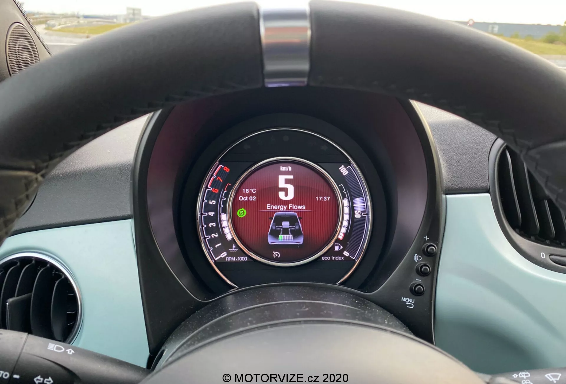 Närbild från förarens perspektiv av interiören i en Fiat 500 2015, med fokus på en lätt sliten svart ratt, en digital instrumentpanel som visar 5 km/h och 'D' för körning, två runda luftventiler och turkosa färgaccenter på instrumentpanelen och dörrarna.