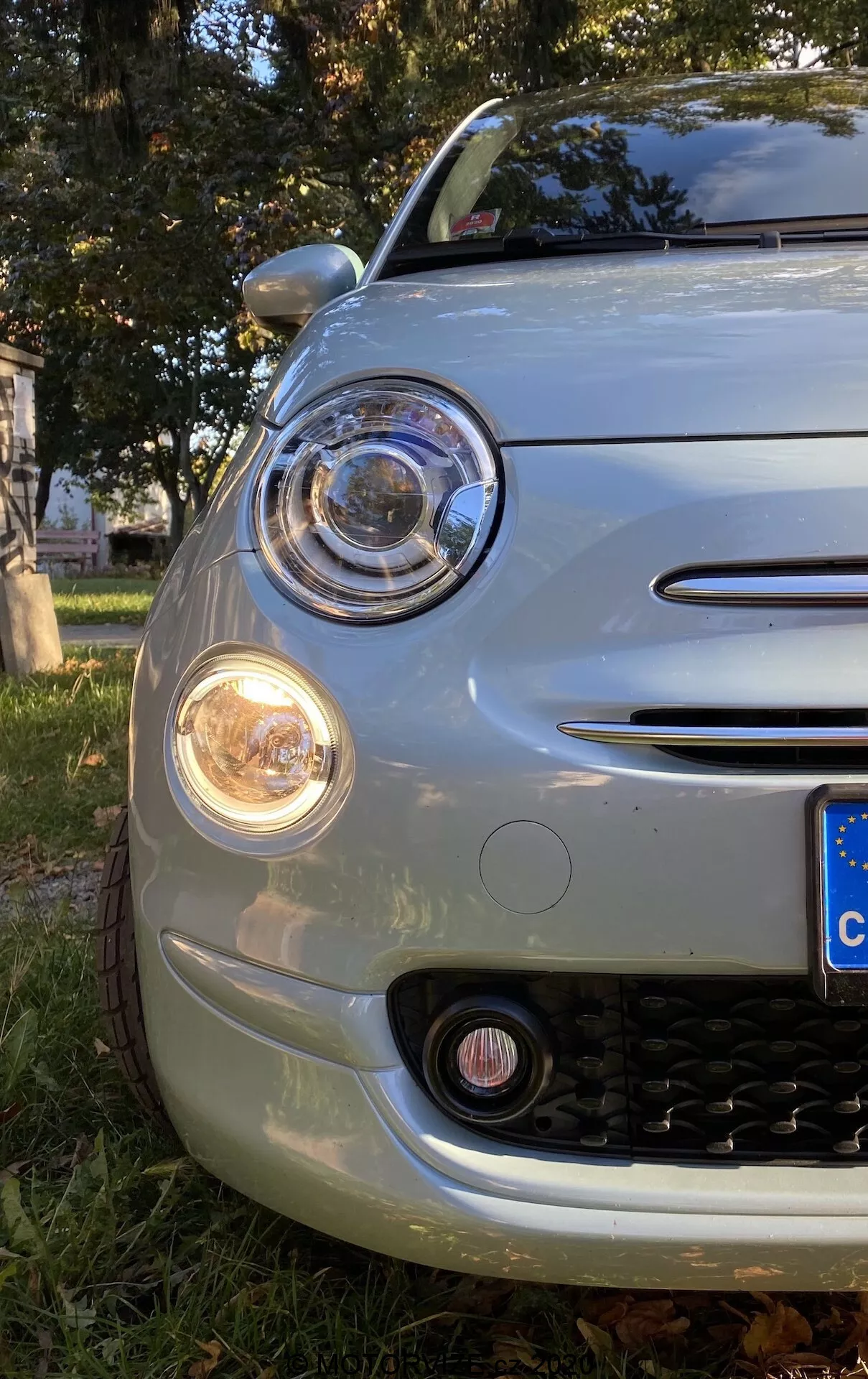 Närbild av vänster framkvartal på en Fiat 500 2015 taget från låg vinkel, med fokus på motorhuv, främre stötfångare, hjulbåge och runt huvudstrålkastare med projektordesign omgiven av en DRL-ring. Signallampan är orange och en dimljus är synlig under. Kromade accenter pryder strålkastarramen, dimljuset och de två horisontella grillribborna. Grillen har ett svart honungskaksmönster. Den rundade fronten och den europeiska registreringsskylten antyder bilens retro-moderna design, parkerad på gräs med höstlöv.