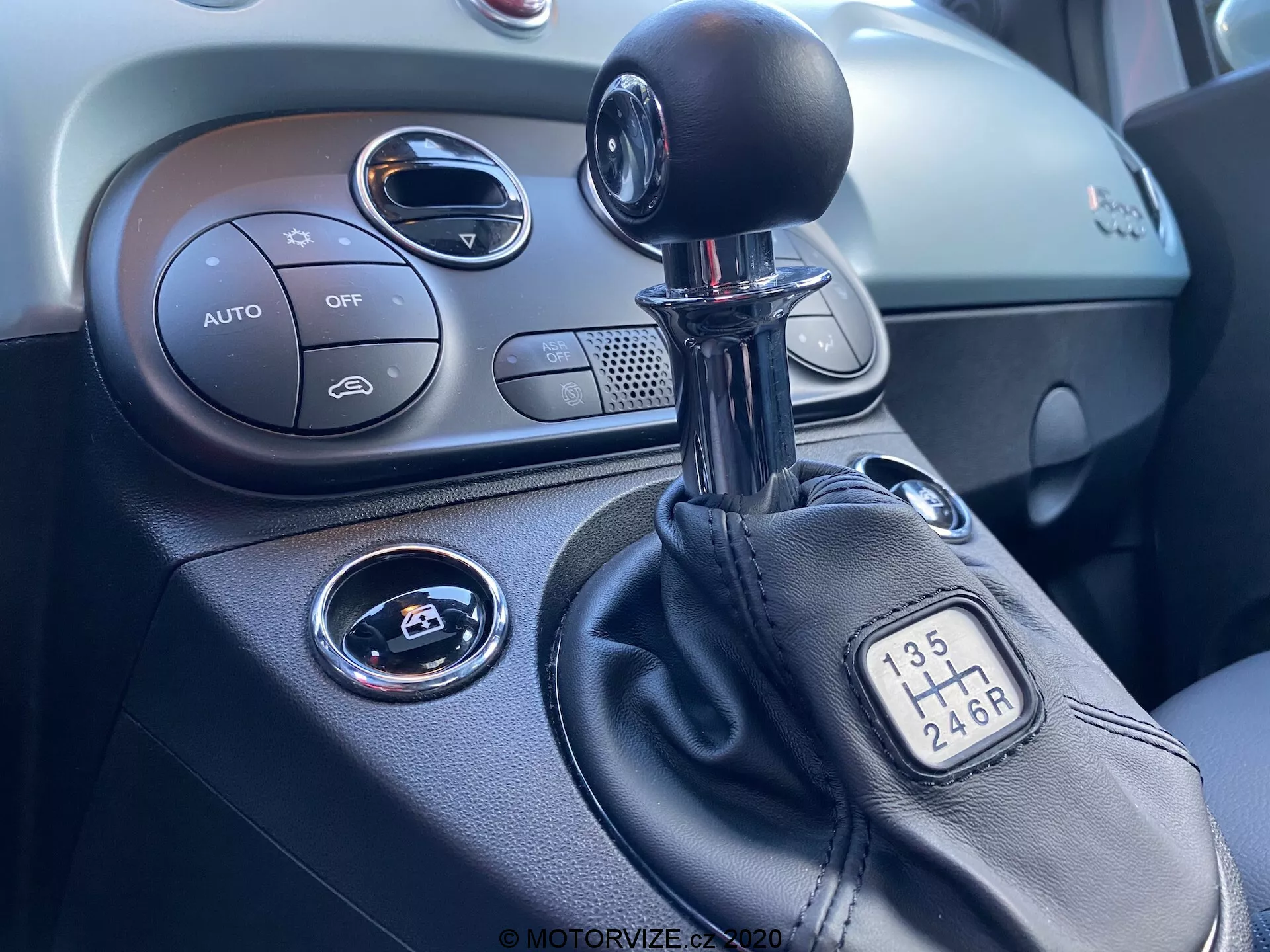Närbild av mittkonsolen inuti en Fiat 500 2015, fokuserad på höger sida. Bilden visar en manuell växelspak med 6-växlad konfiguration, en del av mittkonsolen med klimatkontroller märkta 'AUTO', en 'OFF'-knapp, start/stopp-knappen för tändning, en cirkulär luftventil och den texturerade instrumentpanelen.