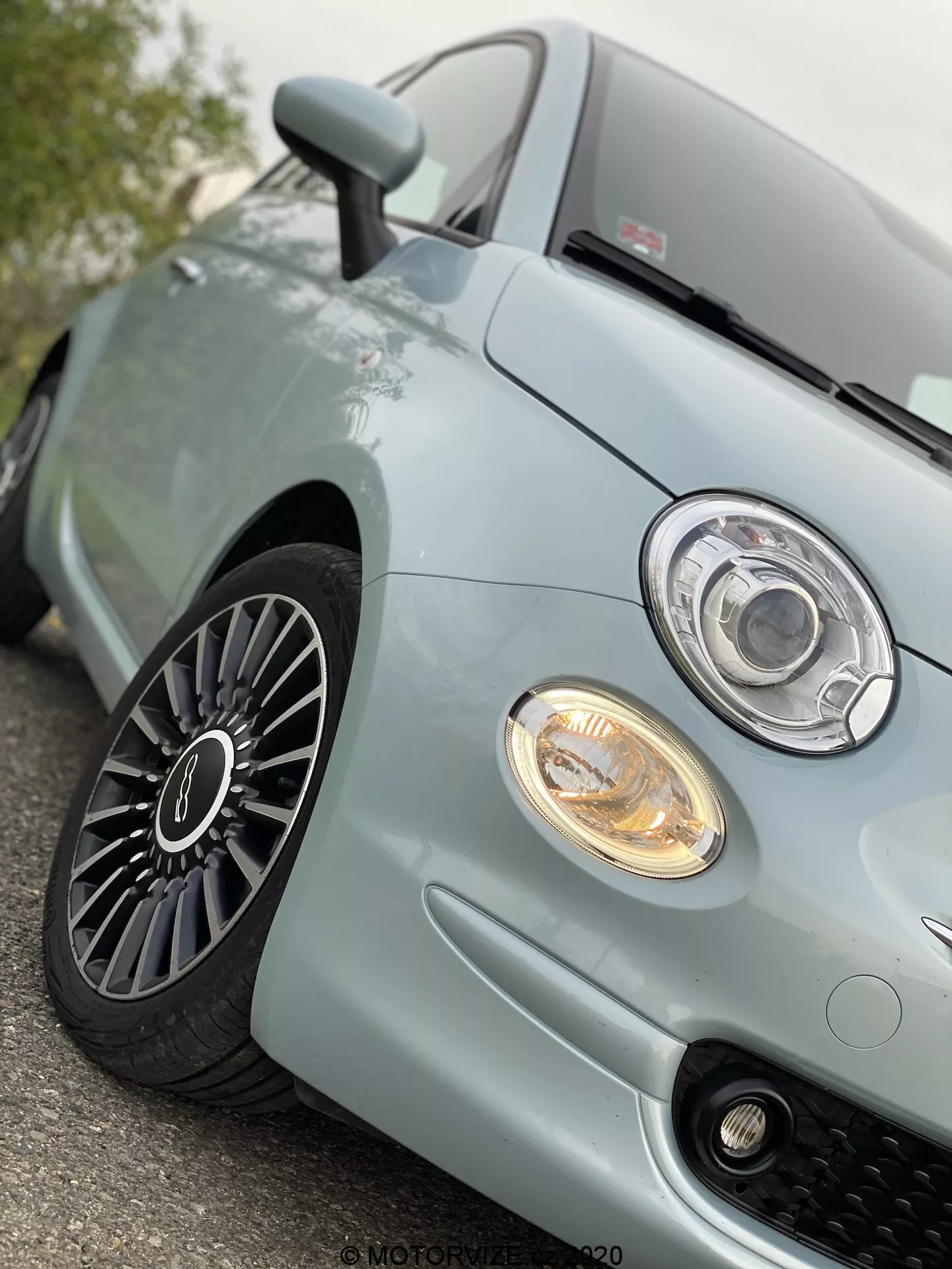 Närbild, låg vinkelvy av vänster framdel på en ljusblå Fiat 500 från 2015, som visar den moderna projektorstrålkastaren med LED- eller halo-ring, belyst dimljus, sportigt honungskakemönstrat grillgaller, främre delen av motorhuven och flerspoke lättmetallfälg med bra slitdjup, vilket antyder en väl underhållen bil och avancerad bildesign.