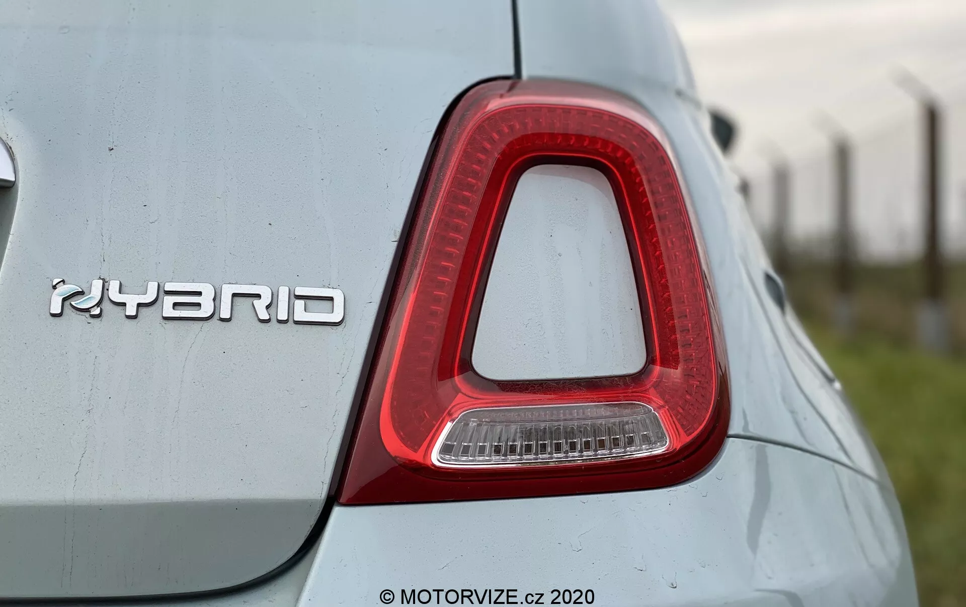 Närbild av den högra baklyktan och 'HYBRID'-märket på en Fiat 500 2015. Baklyktan har en inverterad L-form, främst röd med en klar sektion för backljuset, och är integrerad i bilens kaross. Den metalliska 'HYBRID'-märket, som indikerar hybridteknik, är framträdande placerad. Bilens ljusa färg har en reflekterande kvalitet med några damm- eller vattendroppar, vilket tyder på en utomhusmiljö. Bilden är tagen från en något låg vinkel och framhäver baklyktans design och hybridteknologin.