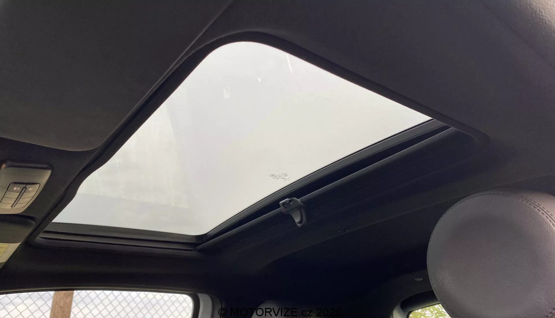 Interiörvy av en Fiat 500 från 2015 med fokus på den övre delen mot taket från en passagerares eller förarens perspektiv. Den framträdande stängda soltaket tillåter naturligt ljus att komma in i kupén. Den mörkfärgade innertaket kontrasterar med exteriören. Synligt handtag på vänster sida, en standard kupolbelysning ovanför och kanterna på två solskydd högst upp. En klädhängare är nära kanten av soltakets glas, som har en säkerhetsetikett längst ner. Bilden är tagen från en låg vinkel, nära inramande takets inre funktioner.