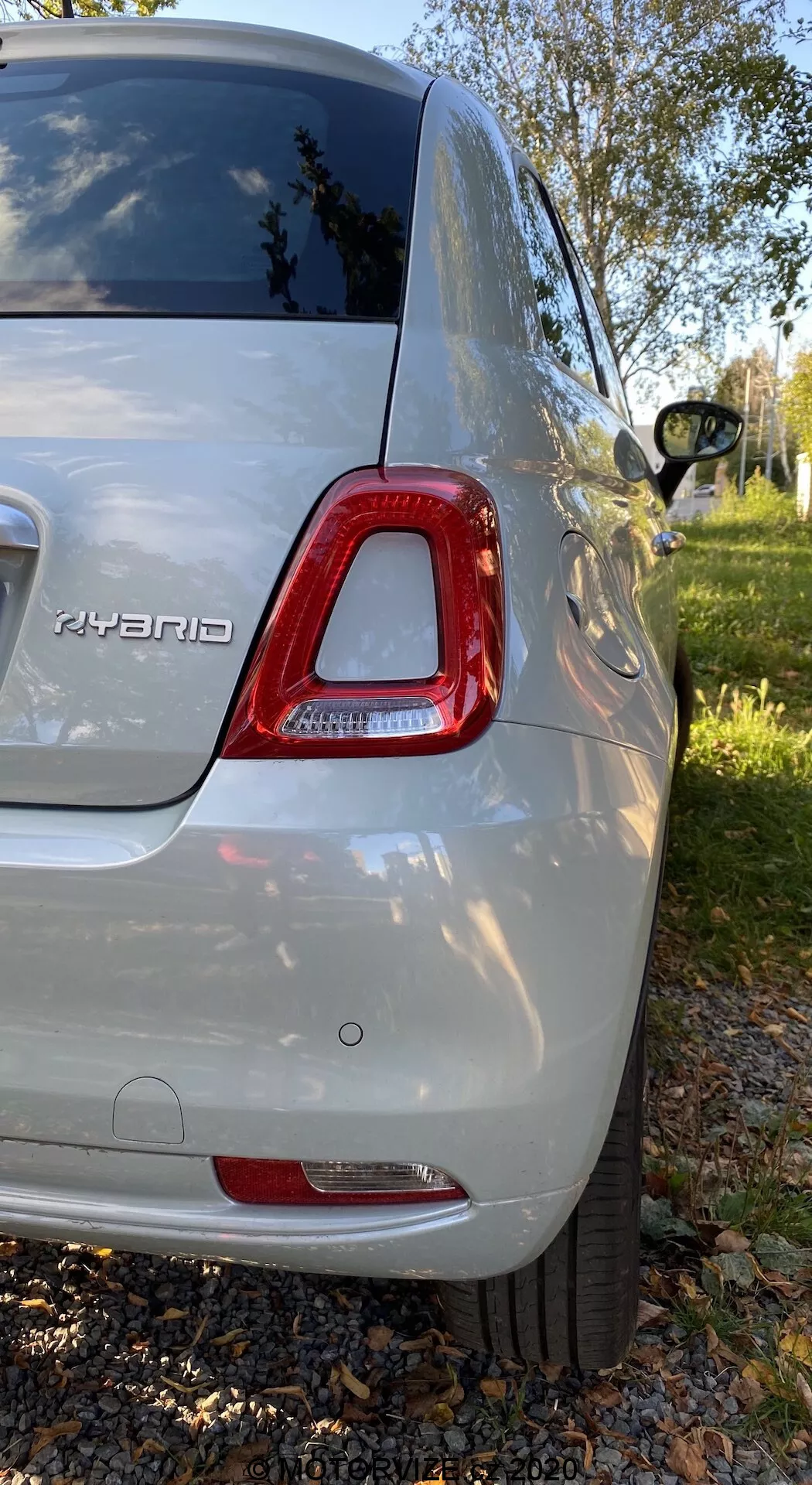 Bakifrån-höger vy av en Fiat 500 hybrid från 2015 i metallic silver, som visar LED-bakljus, hybridmärke, bakre stötfångare, del av legeringshjul, tonade bakre rutor och glänsande kaross; fotograferad i en förortspark med grönska och träd.