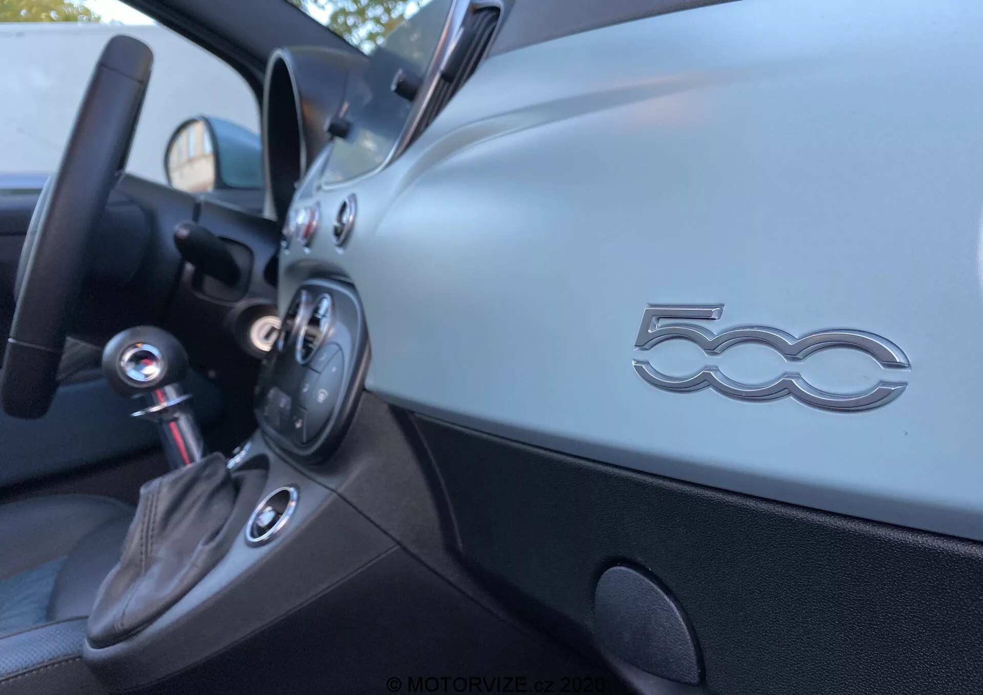 Interiörsbild av en Fiat 500 2015 från framsätet, som visar en sned vinkel av förarsidan av instrumentbrädan. Den matta metalliska finishen på instrumentbrädan har en framträdande '500'-logga, kontrollknappar och en cirkulär ventil. Nedanför finns en svart panel och en växelspak klädd i läder med en kromfinish, vilket tyder på en manuell växellåda. Den inre dörren har ett ergonomiskt handtag och ett förvaringsfack. Fokuset är grunt och lyfter fram '500'-emblemet medan andra element är något suddiga.