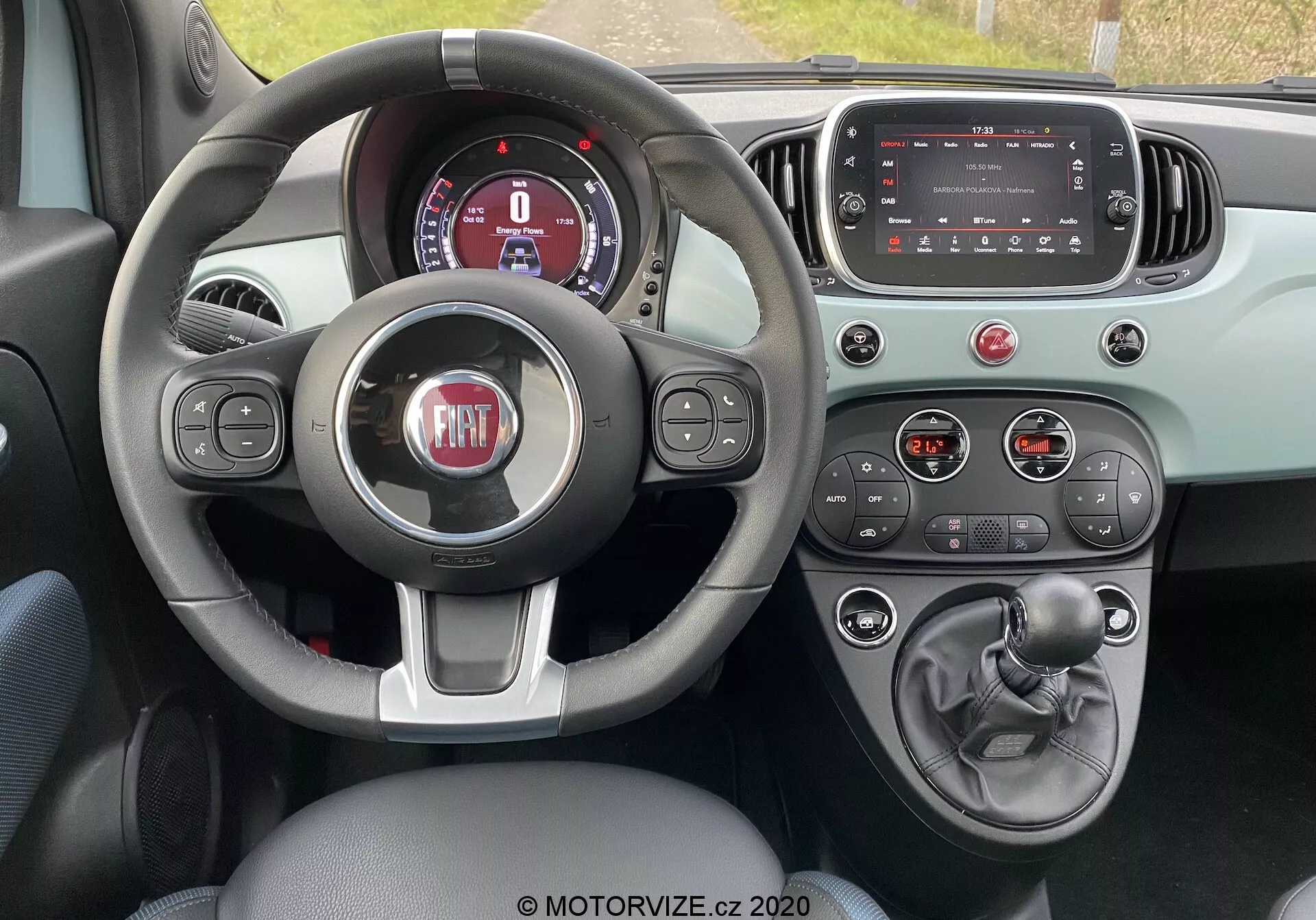 Interiör av en Fiat 500 2015 från förarens perspektiv som visar en tre-ekrad ratt med kontrollknappar, delvis synlig digital instrumentpanel, avstängd infotainment pekskärm, klimatkontrollknappar och vred, manuell växelspak, handbroms och tvåfärgat färgschema som betonar funktionalitet och moderna designelement.