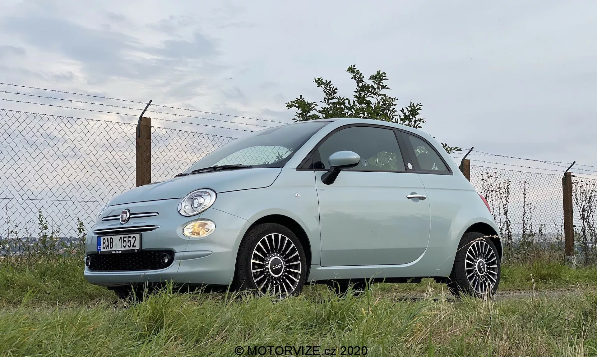 Låg vinkel utomhusfoto av en Fiat 500 från 2015, som framhäver dess robusta hållning. Sidoprofil och delvis framifrån, med runda strålkastare, kromdetaljer och synligt emblem. Tvådörrars pastellfärgat fordon parkerad på gräs med industriell eller semi-urban omgivning. Flerekrade hjul, karossfärgade sidospeglar och synlig bakantenn. Modest främre grill med Fiat-emblem i krom.