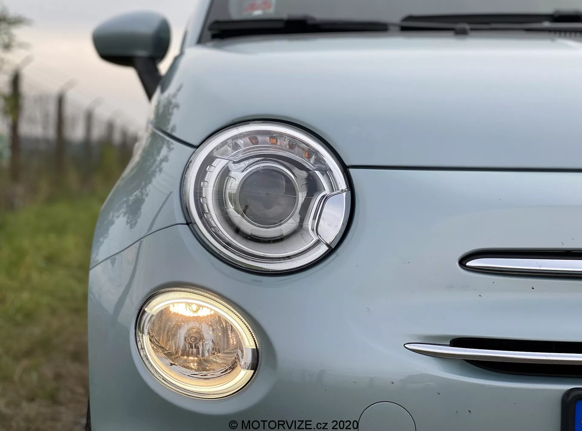 Närbild av en ljusblå Fiat 500 2015 tagen från vänster frontkvartal. Fokus ligger på den distinkta strålkastarensamlingen med cirkulär design och klar lins, samt den mindre cirkulära dimljuset under. Till vänster syns ett segment av grillen med kromade horisontella ribbor. Bilens ljusblå, glänsande lack återspeglar naturligt ljus, vilket framhäver dess välskötta yta. Fordonets märken och logotyper har suddats ut.