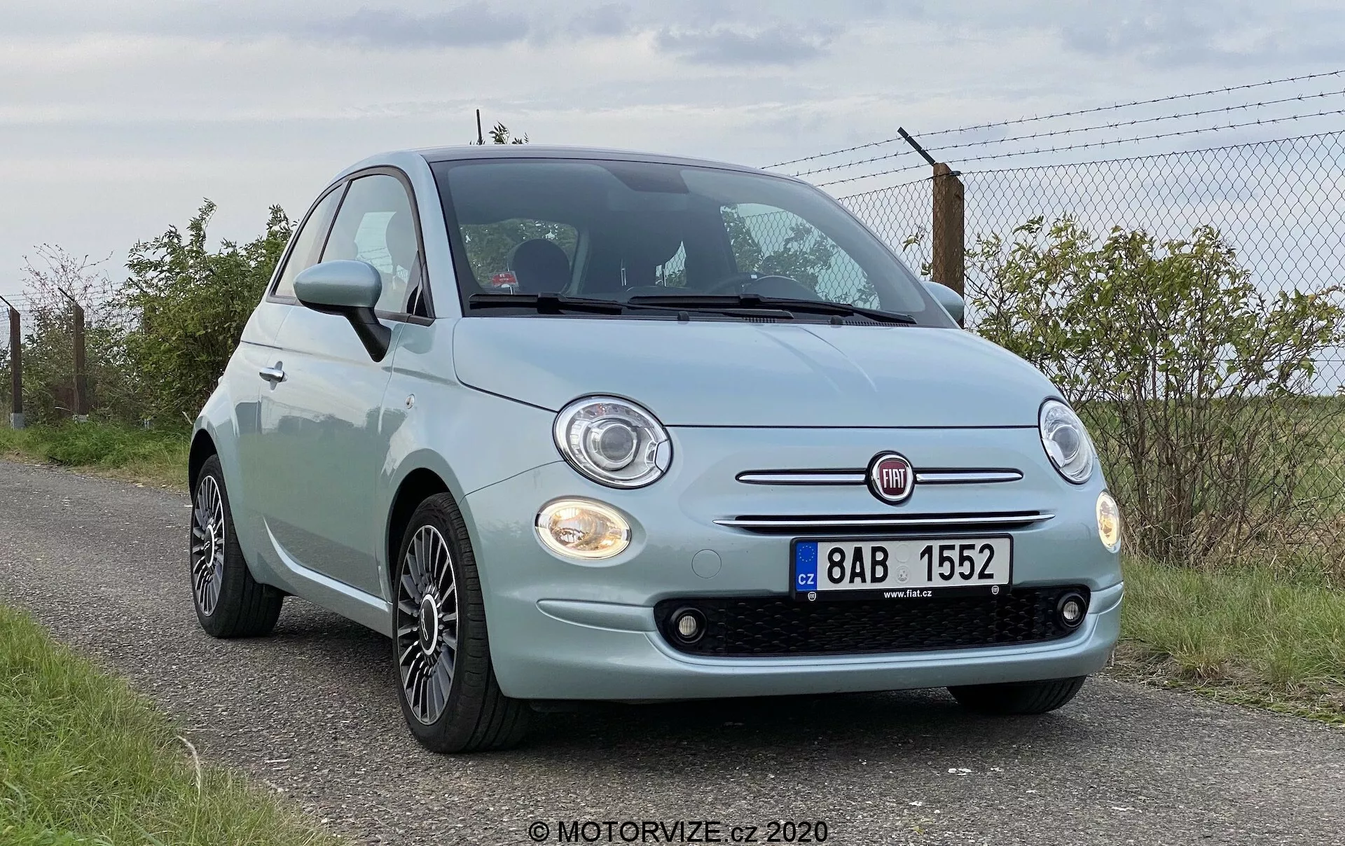 Låg vinkel frontvy av en Fiat 500 från 2015, som visar upp fronten, runda strålkastare, kromdetaljer runt grillen, dimljus och det framträdande Fiat-emblemet. Bilens motorhuv har karakteristiska kurvor och den främre stötfångaren har synliga parkeringssensorer. Även lättmetallfälgar är synliga. Fordonet är parkerat vid vägkanten med en gräsyta och ett staket i bakgrunden, vilket tyder på en lantlig eller halv-lantlig miljö.