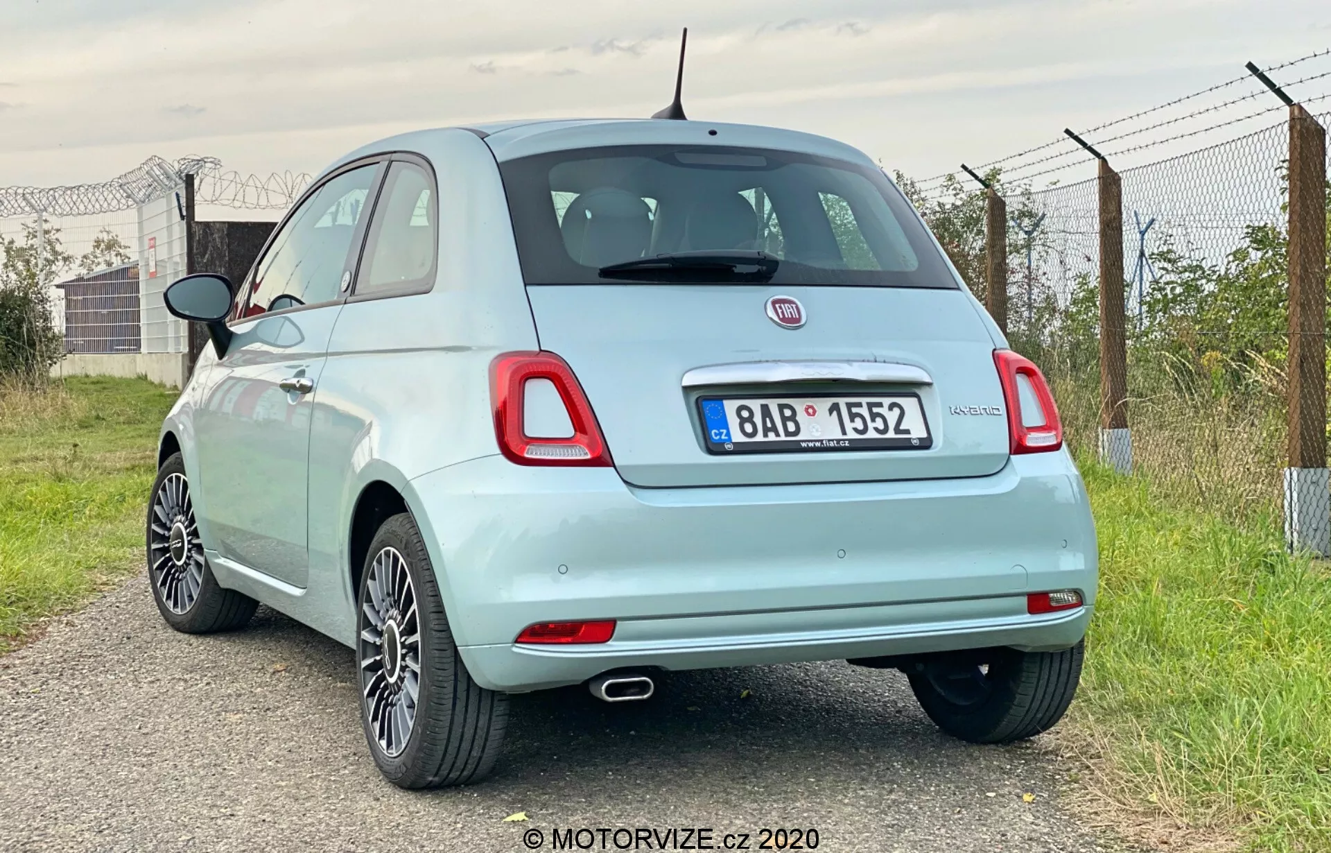 Bildens visar en trekvarts bakåtvinkel på en ljusblå 2015 Fiat 500 halvkombi. Fokus ligger på dess bakdel och del av sidprofilen, tagen från fordonets högra baksida. Den framhäver det högra bakhjulet, bakljuset, delvis vy av högerspegeln och passagerardörren. Bilen är placerad något åt vänster, med kameravinkeln något lägre än taklinjen. Anmärkningsvärda detaljer inkluderar en enkel avgasrör, lättmetallfälgar, standard bakljus, märkes- och modellmarkeringar, en liten bakre spoiler, en maststil antenn och en synlig bränslekork. Miljön verkar vara lantlig med öppna himlar, grönska och ett staket.