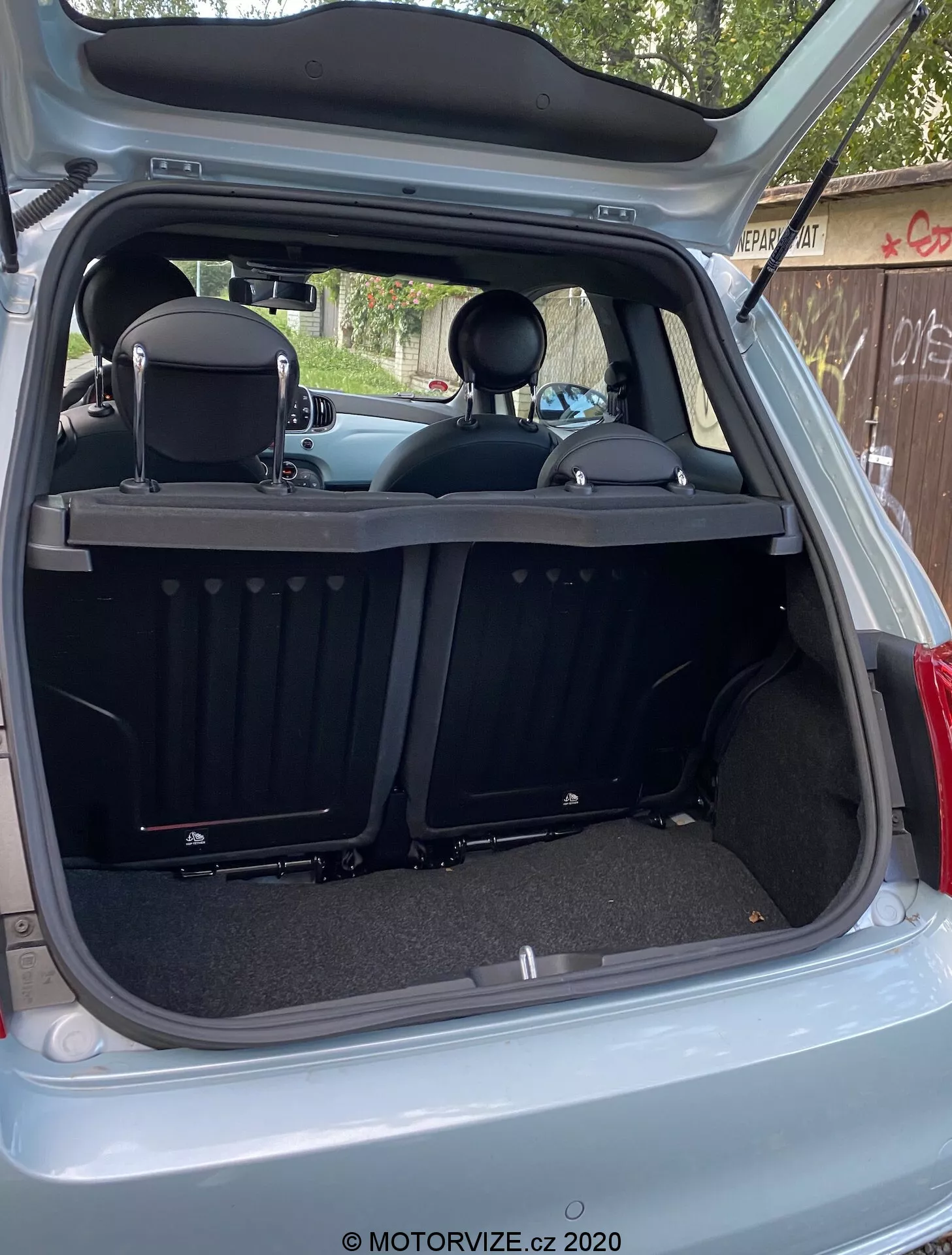 Bilden visar bakre interiören av en Fiat 500 2015 från en yttre, bakre vinkel med bagageluckan helt öppen. Lastutrymmet ses från en något icke-centrerad högersidig vinkel, vilket avslöjar bagageutrymmets djup och olika bakre interiördetaljer. Den moderna, rena designen inkluderar mattbelagda ytor, ett infällbart ribbat lastskydd, delbart fällbart baksäte med nackstöd och en skyddslist. Teknologiska funktioner inkluderar en interiörbelysning, ett 12V-uttag, plastmouldningar, runda utskärningar och sidopaneler.