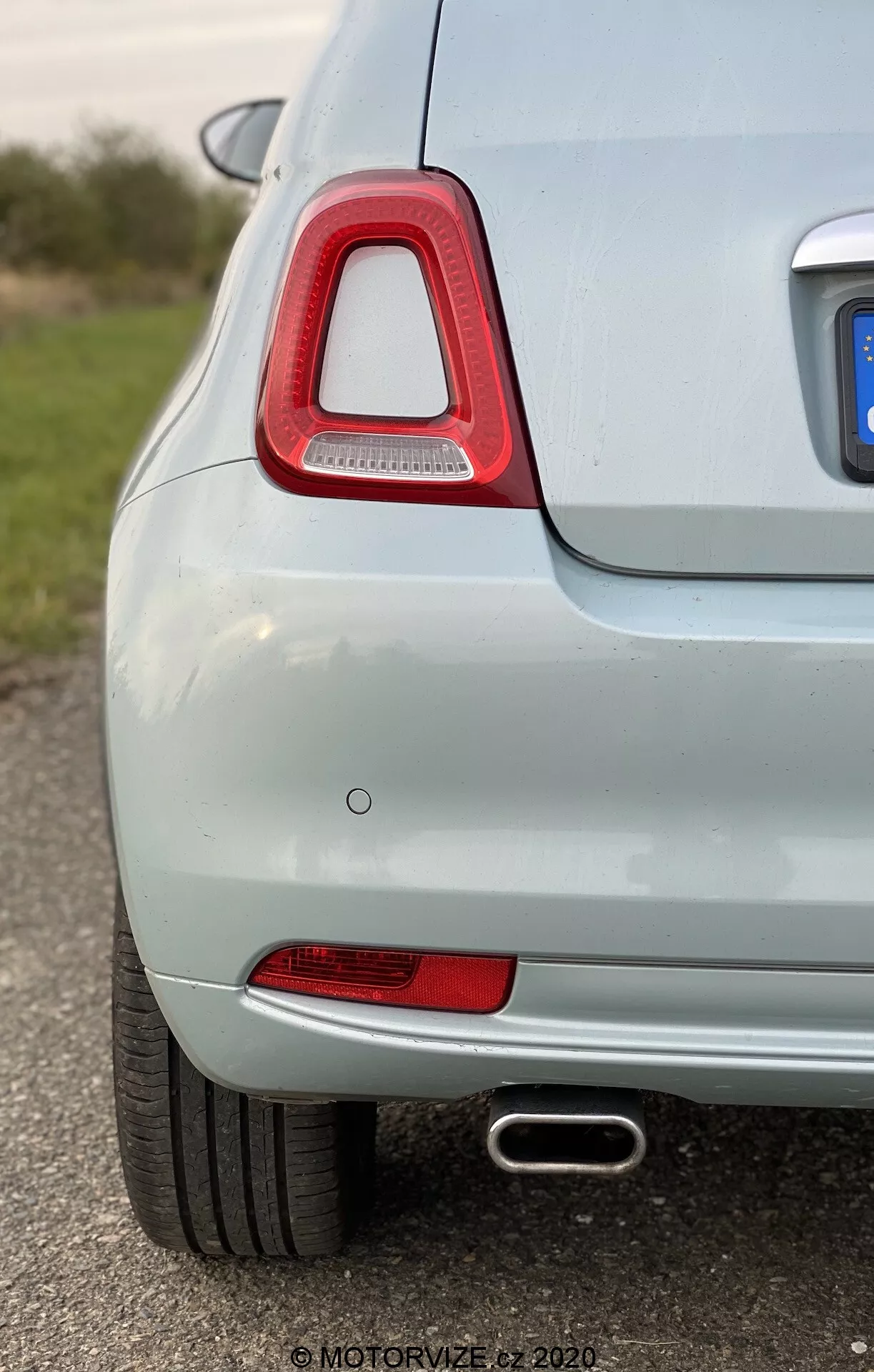 En närbild, sned vy på den bakre vänstra sidan av en ljusfärgad bil, som lyfter fram den distinkta baklyktan med klar röd lins och LED-teknik, röd reflektor, infälld parkeringssensor och kromad avgasrörsdetalj. Den ljusa metallic-lackeringen och det grunda skärpedjupet framhäver fordonets rena och väl underhållna tillstånd.
