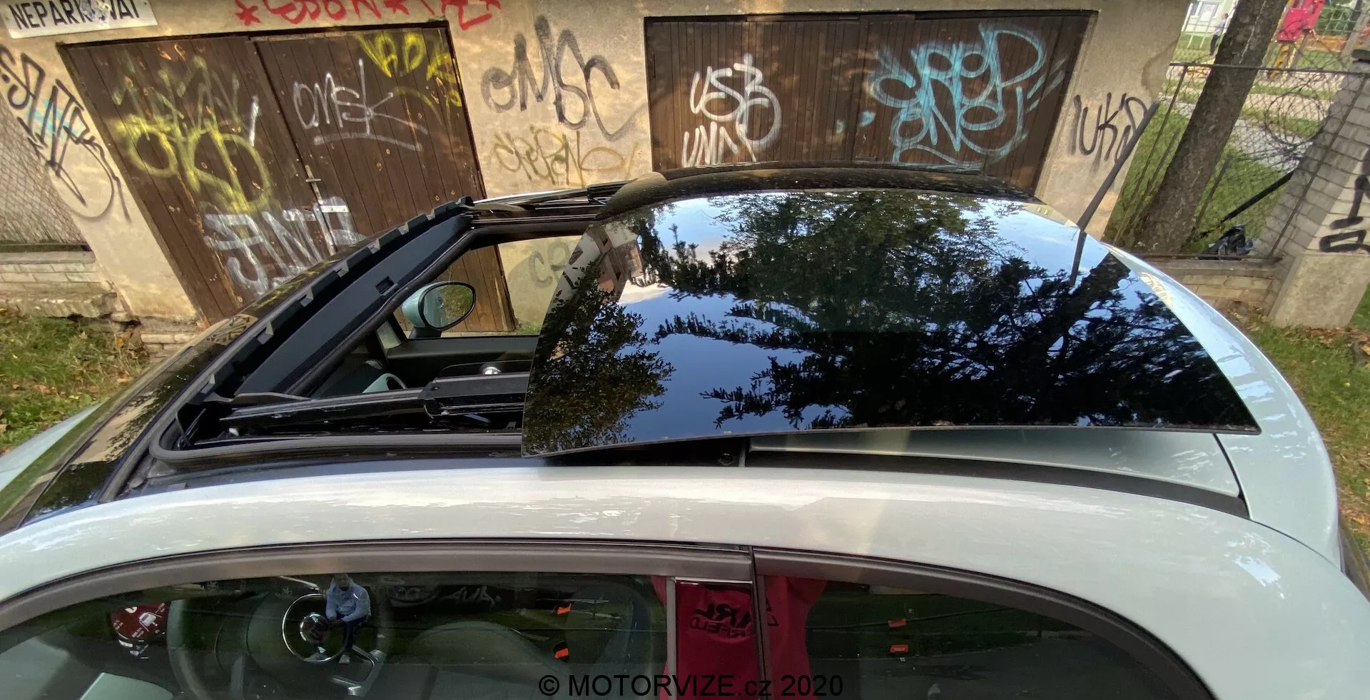 Närbild av bakdelen på en Fiat 500 2015 med fokus på den övre delen, inklusive den stora bakrutan med toning som omsluter sidorna, en sportigare spoiler, ett taklucka/panoramatak, och en öppen baklucka, som reflekterar utomhusgrönska och dagsljushimlen.