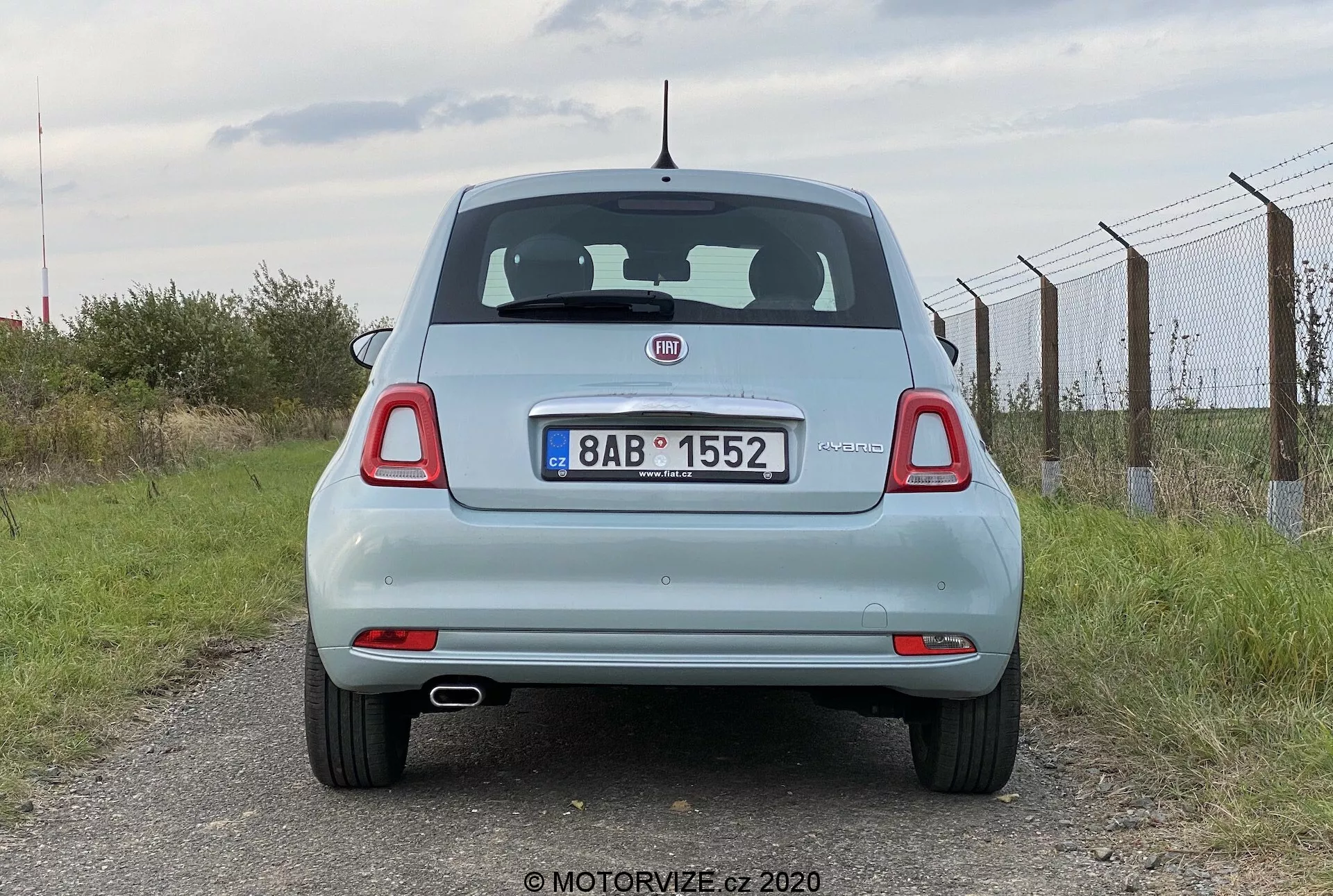 Bakifrån av en ljusblå Fiat 500 2015. Perspektivet i ögonhöjd fångar fordonet som står stilla, och visar dess bakdel inklusive bagagelucka, bakruta, bakljus, bakre stötfångare och registreringsskylt. Funktionerna inkluderar ett framträdande tillverkarens emblem, bakrutespolare, högt placerat bromsljus och runda bakljus med ytterligare segment. Registreringsskylten är monterad i mitten av bagageluckan, under emblemet och ovanför den platta bakre stötfångaren med integrerade reflexer. En kort antenn är placerad mitt på taket mot baksidan. Molnig himmel och lantlig omgivning med ett staket till höger och gräs nedan.