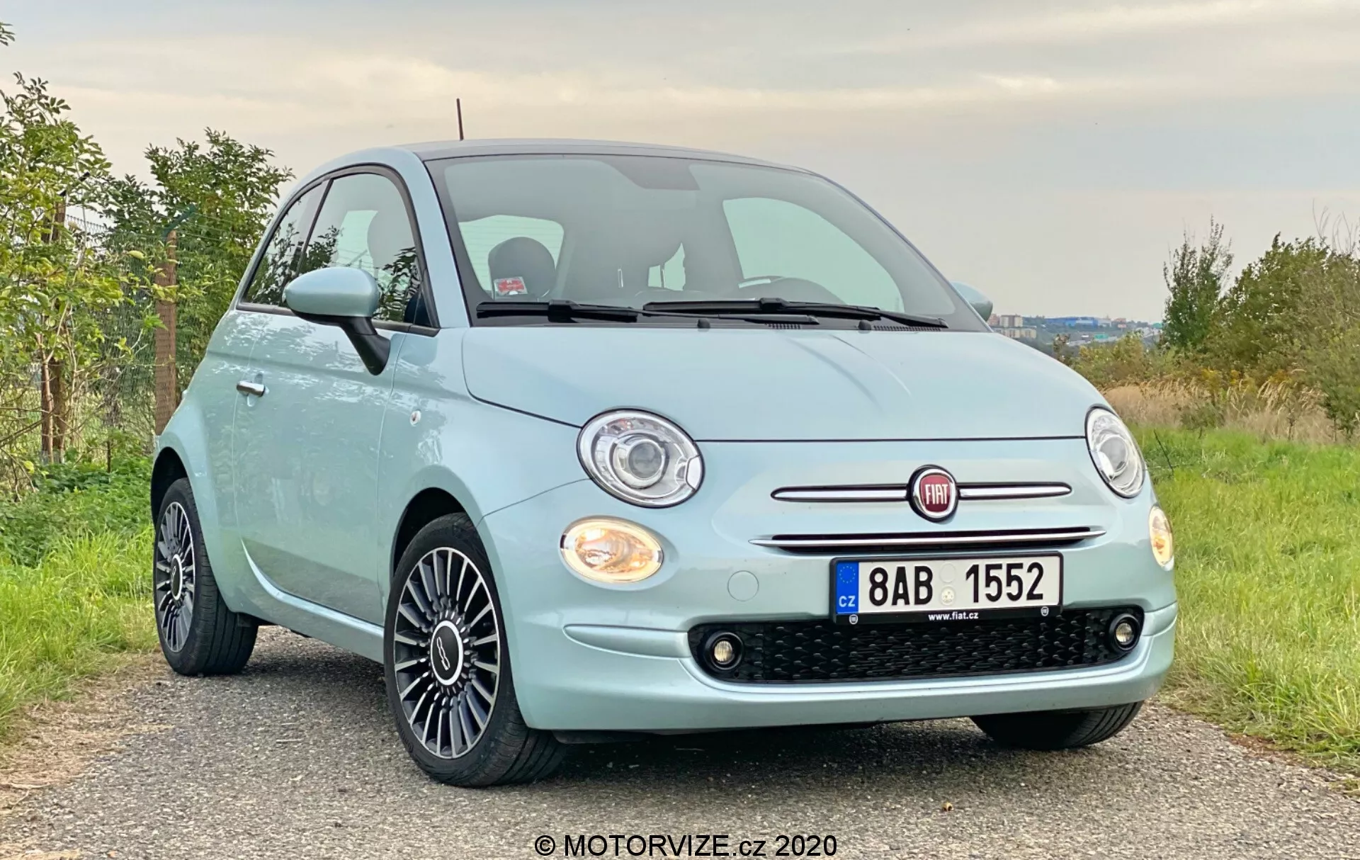 Framifrån i trekvartsvy av en ljusblå Fiat 500 från 2015 med kromad grill och runda halogenstrålkastare vid en något låg vinkel. Bilden framhäver fronten, grillen, strålkastarna, huven och legeringshjulen med svart multipekdesign. Bilen är parkerad på en grus- eller jordväg i ett lantligt område med grönska i bakgrunden.
