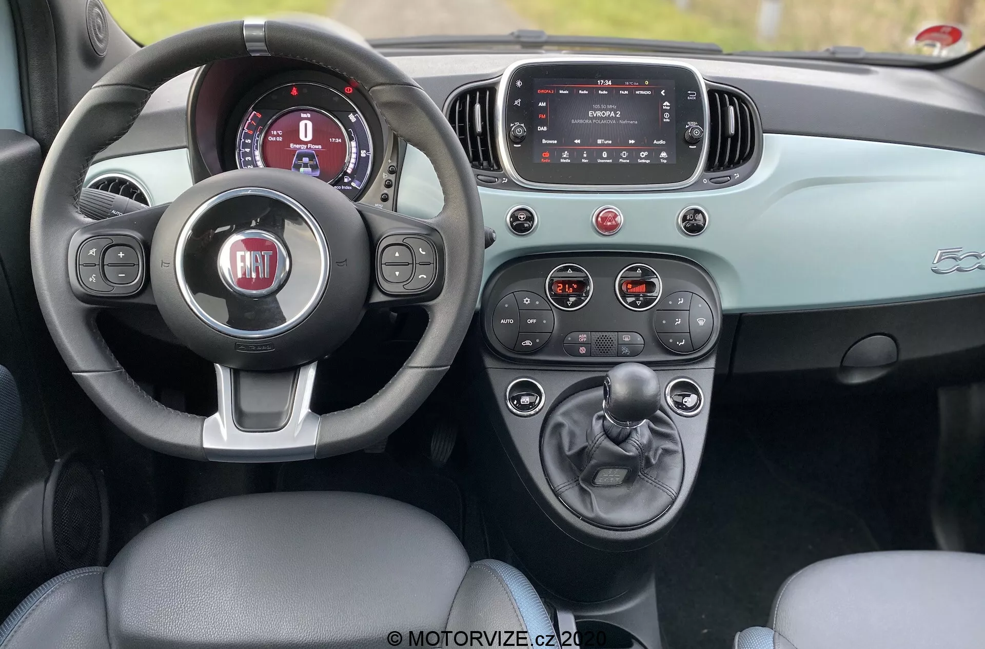 Interiörbild av en Fiat 500 från 2015 sedd från baksätet, som visar förarutrymmet med ett lädersäte, treekrad ratt med FIAT-logotypen, digital instrumentpanel, infotainment-pekskärm och mittkonsol med manuell växelspak och mugghållare. Instrumentpanelen och dörren har ett kontrasterande tvåfärgat färgschema som förbättrar den estetiska designen. Bilden är tagen under dagen och framhäver bilens raffinerade interiördesign och funktionalitet.