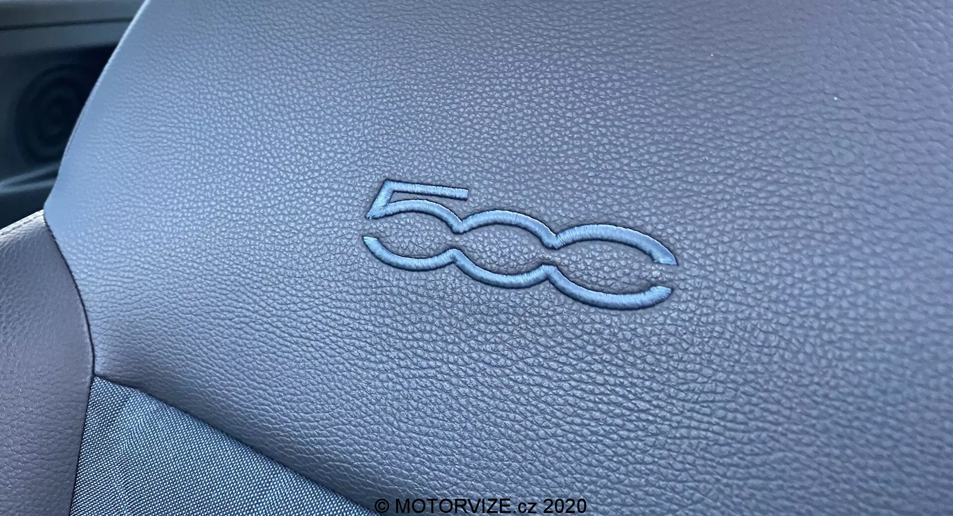 Närbild som visar den stilfullt präglade '500'-logotypen på instrumentpanelen i en Fiat 500 2015 interiör. Den läderliknande texturen och de exakta sömmarna på instrumentpanelens panel framhävs av belysningen och det smala skärpedjupet, vilket betonar den högkvalitativa hantverkskonsten i fordons interiördesign.