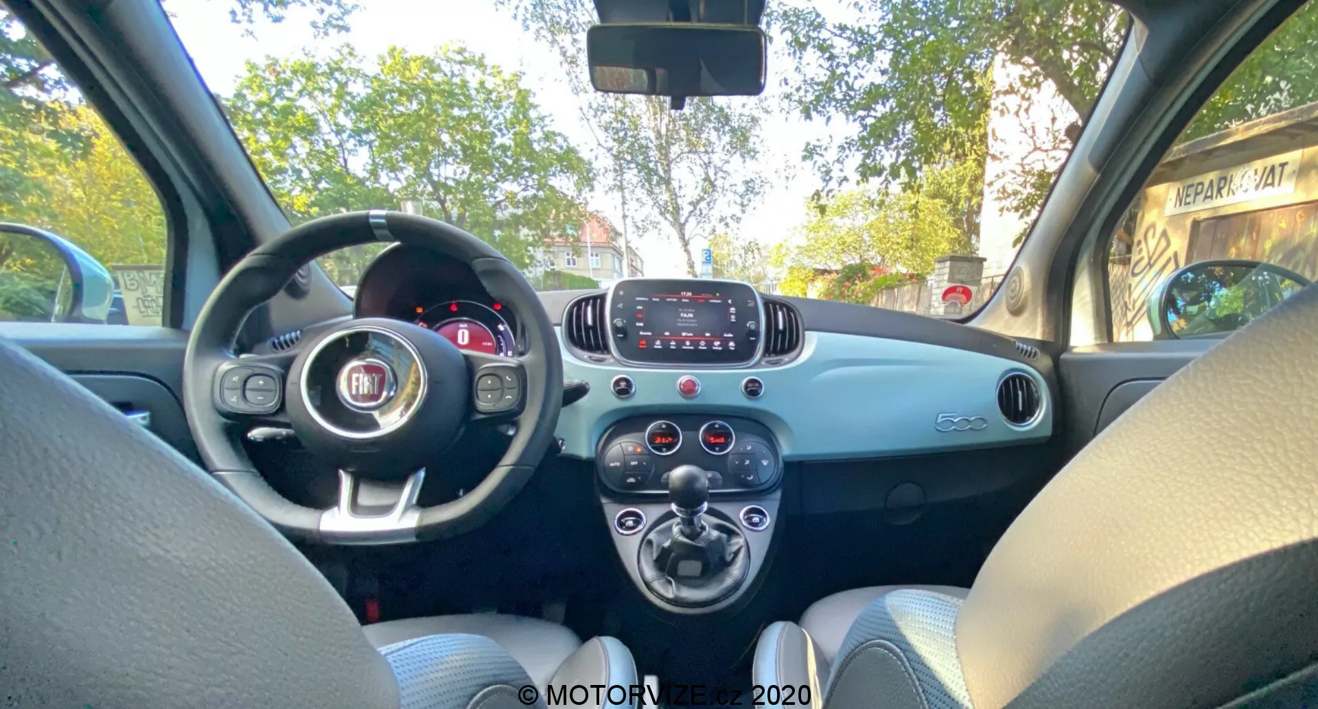 Interiörbild av Fiat 500 2015 från baksätet, som visar framinstrumentbrädan, ratt med Fiat-emblem, delvis synligt instrumentkluster, grundläggande infotainmentsystem med knappar, klimatanläggningsvred, tvåfärgad instrumentbräda, ljusgrå tygsäten, passagerarsidsdörr med handtag och högtalargaller, enkel och funktionell design utan avancerade tekniska funktioner.
