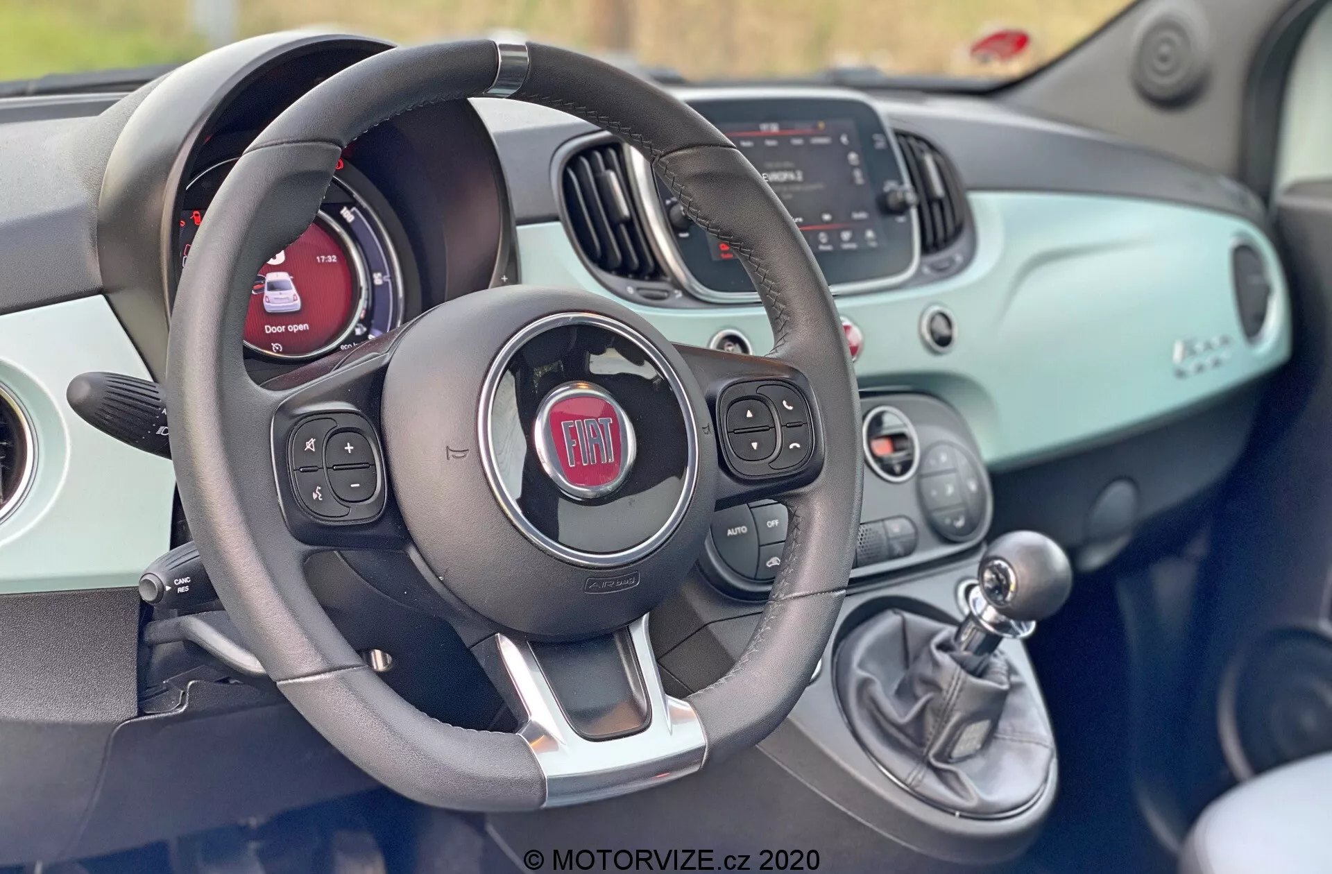 Interiörvy av en Fiat 500 från 2015 från förarsätet, som visar ratten med inbyggda kontroller för ljud, röstkommandon och möjligen farthållare. Bakom ratten finns instrumentpanelen med en digital skärm och traditionella mätare. Instrumentbrädan har en yta i samma färg som karossen och inkluderar en infotainmentskärm som visar information om radio och media samt manuella HVAC-kontroller. Till höger om ratten finns en manuell växelspak som visar på en manuell växellåda. Den övergripande designen är modern med en sportig, tvåfärgad estetik.