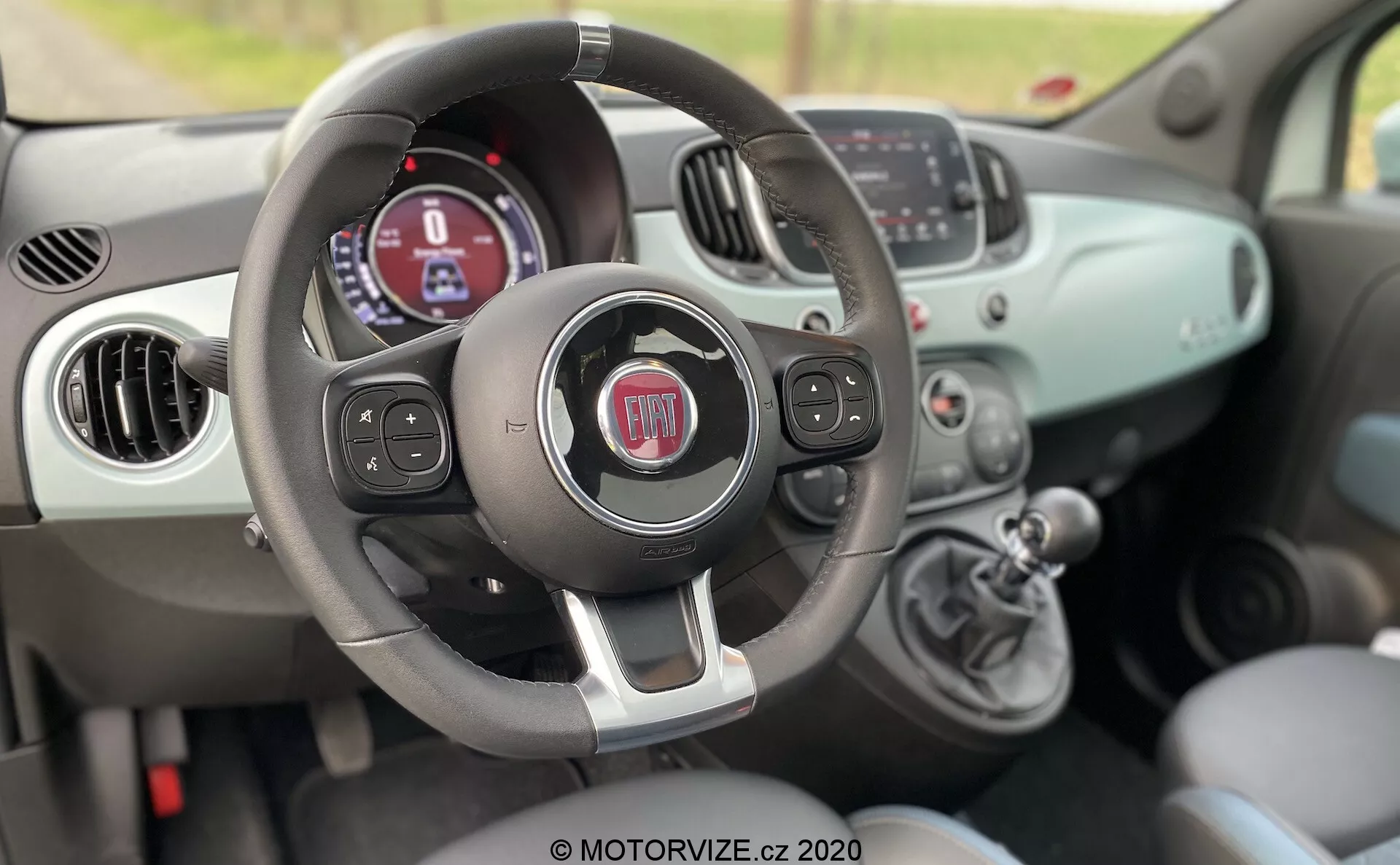 Interiörvy av en Fiat 500 från 2015 från passagerarsidan, som visar ratten med multifunktionsknappar, instrumentpanelen med digital display, manuella klimatreglage och växelspak.
