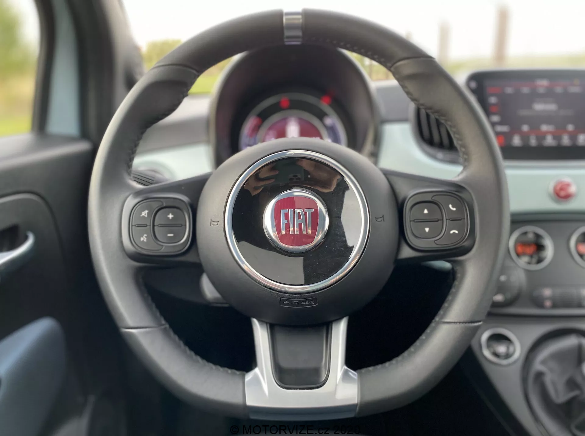 Interiörvy av en Fiat 500 2015 från förarsätet, som visar en modern treekrad ratt med kromdetaljer och Fiat-logotypen i mitten. Ratten har inbyggda kontroller för media-, röstfunktioner och farthållare. Den suddiga bakgrunden innehåller instrumentpanel och pekskärmsinfotainmentsystem, vilket framhäver fordonets samtida teknik och design.