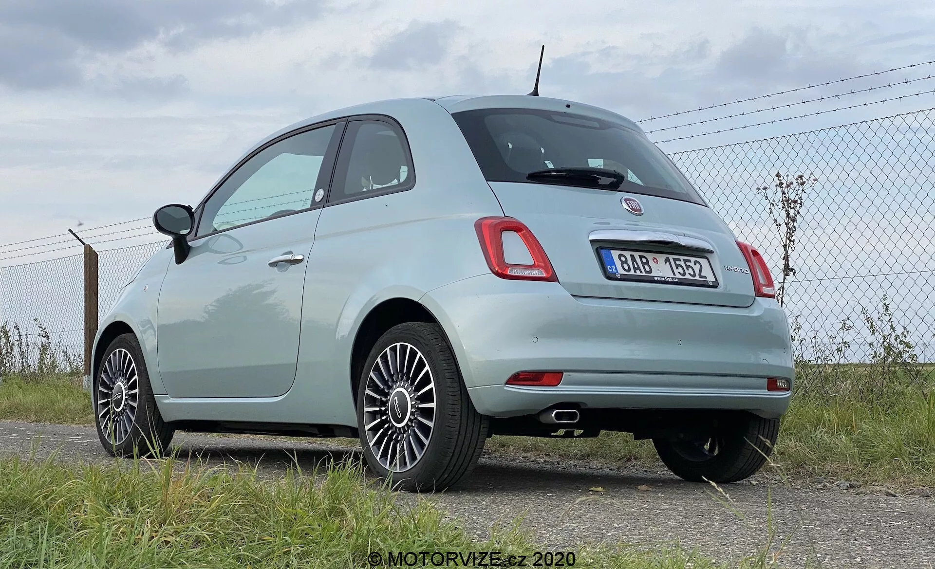Vy bakifrån i tre fjärdedelar av en ljusfärgad modern Fiat 500 hatchback, som visar distinkta runda baklyktor och ett centralt kromemblem ovanför det bakre registreringsskyltsområdet. Bilden framhäver fordonets rundade taklinje, kromlister runt sidorutorna och multiekrade aluminiumhjul. Bilens ljusa pastellfärg indikerar att den är relativt ny eller väl underhållen. Den är parkerad nära ett kedjelänkstaket på ett asfalterat område, med himlen och horisonten synliga i bakgrunden, vilket antyder en flygplats eller öppen mark. Möjliga tekniska funktioner inkluderar bakre parkeringssensorer integrerade i den svarta delen av bakre stötfångaren.