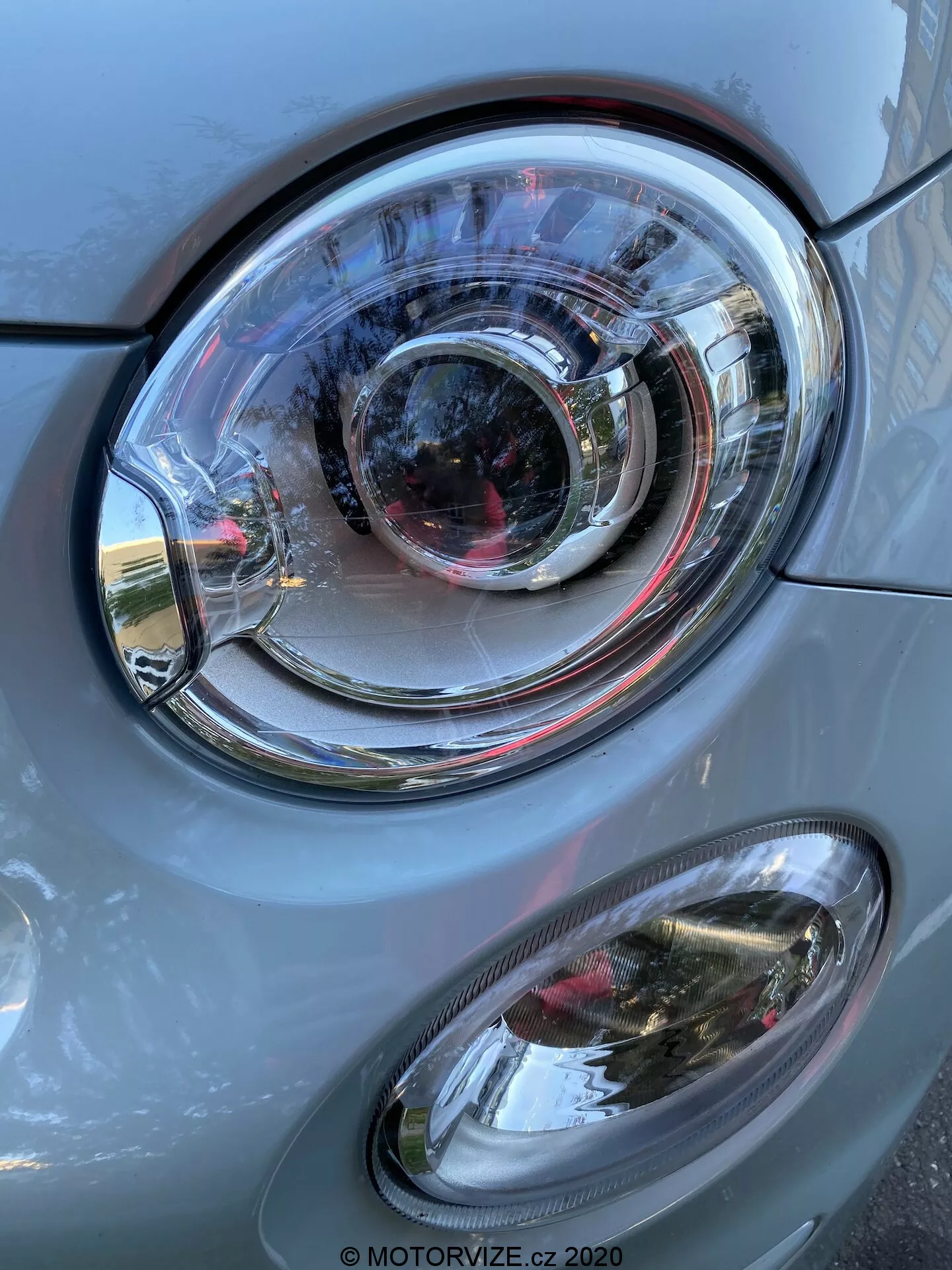 Närbild av framsidan på en Fiat 500 från 2015, med fokus på strålkastaren. Bilden, i vertikalt orientering, visar en framstående huvudstrålkastare med en klar polykarbonatlins och en komplex design av koncentriska ringar, vilket antyder en projektorlampa och eventuellt adaptiv belysningsteknik. Nedan finns en sekundär dimljus med oval form och transparent hölje. Karossens grå färg och metalliska glans har en slät yta, som reflekterar dagsljuset och omgivande element såsom byggnader och himmel. Strålkastaren och dimljuset ser ut att vara i gott skick, vilket tyder på en väl underhållen eller ny bil.
