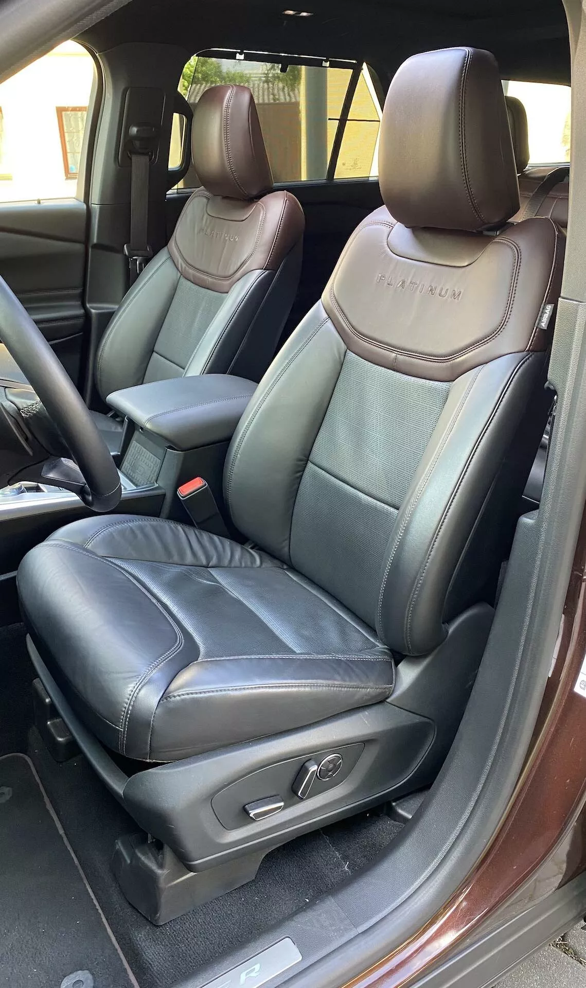 En detaljerad vy av interiören i en 2019 Ford Explorer, med fokus på förarsätet med den öppna dörren. Fotografiet framhäver det svarta läderklädseln med kontrasterande sömmar, en präglad logotyp på sätesryggen, justerbart nackstöd, säteskontrollknappar, mittkonsolens armstöd, synligt bältesfrigöringsknapp, intern dörrlåsning och metalliska dörrtröskelkanter.