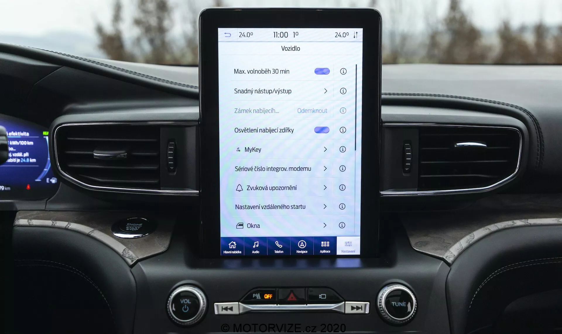 Närbild av interiören på en 2019 Ford Explorer med fokus på mittkonsolen och infotainmentsystemet. Den vertikalt orienterade pekskärmen visar ikoner och texter som antyder funktioner som navigation, fordonsinställningar, mediekontroller, Bluetooth och möjligen Apple CarPlay eller Android Auto. Instrumentpanelen inkluderar vertikala luftventiler, ambient belysning, fysiska knappar och vred för klimat- och volymkontroll. Övre delen av en multifunktionsratt och en delvis digital instrumentpanel som visar fordonsinformation är också synliga. Designen framhäver ett modernt fordon i mellanklass eller premiumklass med en mörk färgpalett.