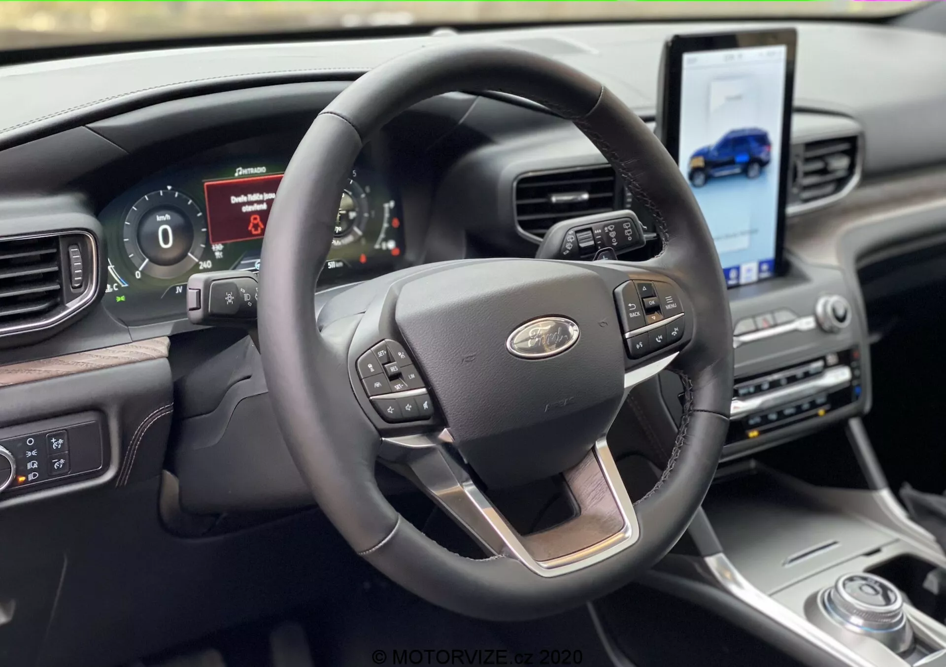 Interiör vy av en Ford Explorer 2019 från förarsätets perspektiv, med en modern tre-ekrad ratt med kromdetaljer och multifunktionsknappar för farthållare, media och röstkommandon. Bakom ratten finns en digital instrumentpanel som visar fordonsinformation. Instrumentbrädan inkluderar en stor pekskärm vinklad mot föraren för navigering och medieåtkomst, kompletterad med en roterande växelväljare och exklusiva material.