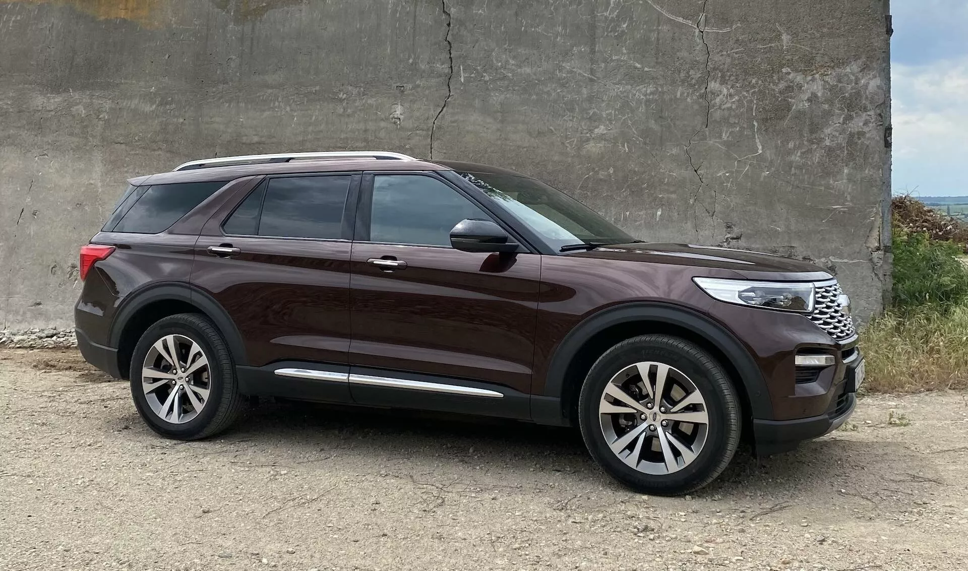 2019 Ford Explorer SUV parkerad på grov grus nära en betongstruktur, sidoprofilvinkel som framhäver designelement som stora legeringshjul, hög linje, tonade fönster, karossfärgade dörrhandtag, kromremsa vid fönster, snygga karosslinjer, integrerade blinkers i dörrspeglar, djup metallisk lack och märkbar markfrigång.