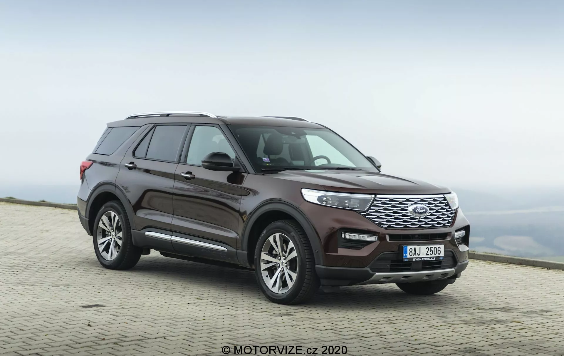 Bild av en 2019 års Ford Explorer SUV tagen från en tre-kvarts frontvinkel, som framhäver både sidoprofilen och fronten. Den låga kameravinkeln betonar fordonets robusta hållning och framträdande frontgrill. Bilens hela längd är synlig, visande desiglinjer, hjuldesign och konturerna av dörrarna och skärmarna. Hjulens är något vridna åt höger, vilket ger en känsla av rörelse. Framträdande drag inkluderar en frontgrill med horisontella ribbor, eleganta strålkastare som integreras med grillen och en främre stötfångare med synliga sensorer. Sidospeglarna verkar inneha integrerade blinkers, och taklinjen lutar något bakåt med synliga takräcken. Lackeringen är i en djup metallisk vinröd färg. Fordonet är parkerat på en asfalterad yta med en suddig naturlig bakgrund, vilket antyder en hög höjd med en molnig himmel.