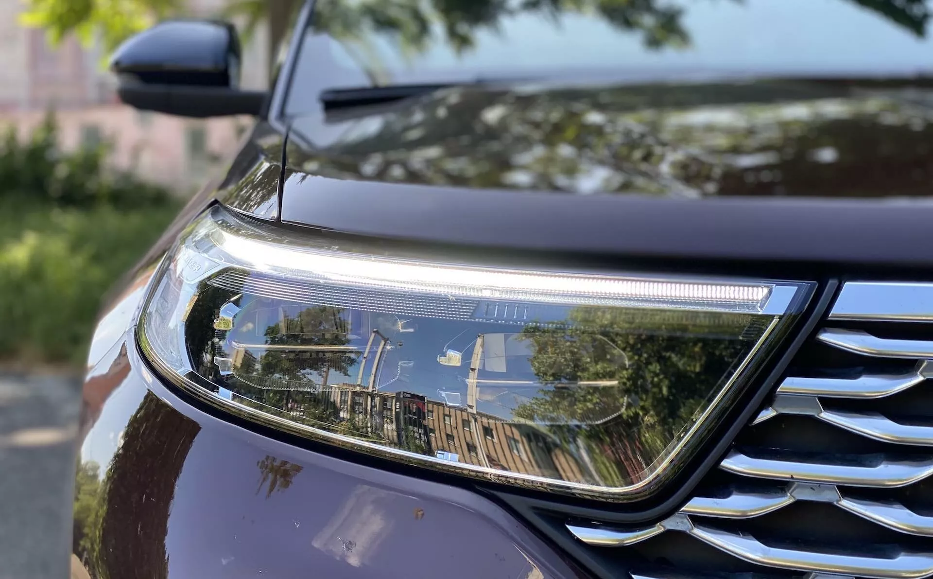 Närbild av främre delen av 2019 års Ford Explorer som visar höger strålkastare och en del av grillen. Strålkastaren har en modern LED- eller HID-design med en horisontell DRL, medan grillen har kromaccenter med horisontella ribbor. Bilens mörka, metalliska lack reflekterar omgivningen och framhäver utomhusinställningen. Bilden fångar också en del av skärmen och huven och betonar aerodynamiska konturer.