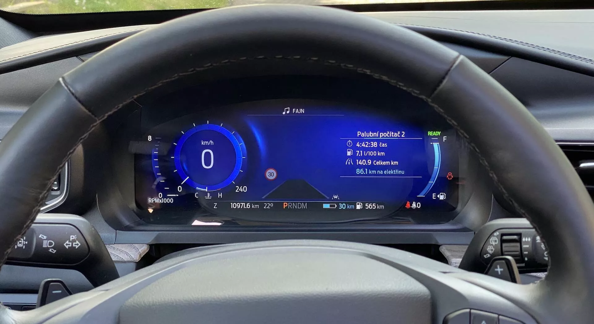 Interiörvy av en 2019 Ford Explorer från förarens perspektiv, med fokus på det centralt placerade ratten och den digitala instrumentpanelen bakom den. Ratten har olika kontrollknappar på båda ekrarna. Bakom den visar en helt digital instrumentpanel information som hastighet, effektmätare, körsträcka, bränslenivå, utomhustemperatur och förbrukningsavläsning. Instrumentpanelen visar också tid och datum och är designad med moderna, teknologiskt avancerade element i blått och vitt. Ratten är gjord av svart läder med sömdetaljer.