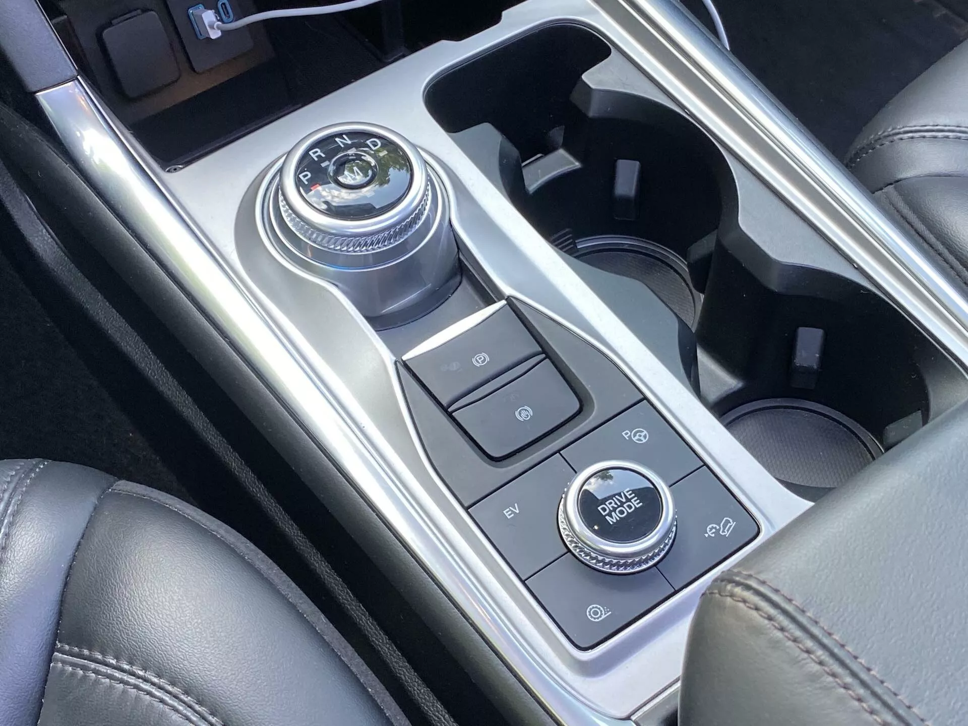 Närbild av mittenkonsolen i en Ford Explorer 2019 med en roterande växelvred, olika kontrollknappar, mugghållare och en infotainmentsystemvred, allt omgivet av högblank metallic och matta sektioner som antyder modern lyx och avancerad teknik.