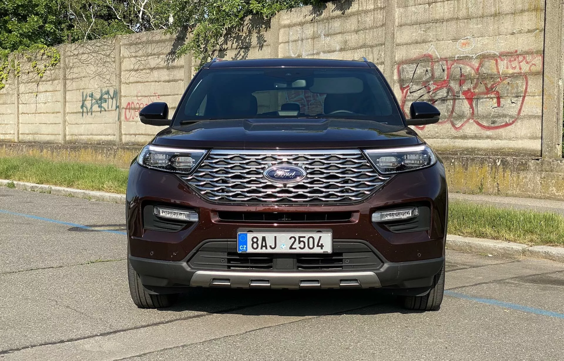 Frontal vy av en 2019 Ford Explorer SUV fångad från en vågrät vinkel, som visar fordonets moderna design. Den framträdande fronten har ett stort, kromat galler med hexagonalt mönster och tillverkarens emblem centralt placerat. Eleganta, horisontellt förlängda LED-strålkastare och en färgmatchad främre stötfångare med nedre luftintag och parkeringssensorer framhäver teknologiska framsteg. Huvens konturerade design med upphöjda linjer och skärmarna samt de integrerade blinkers i sidospeglarna ger ett muskulöst utseende. SUV:n, målad i en rik kastanjebrun eller mörkröd färg, är parkerad på en asfalterad yta med en betongkant och en graffitibeklädd vägg i bakgrunden, vilket framhäver en urban miljö.