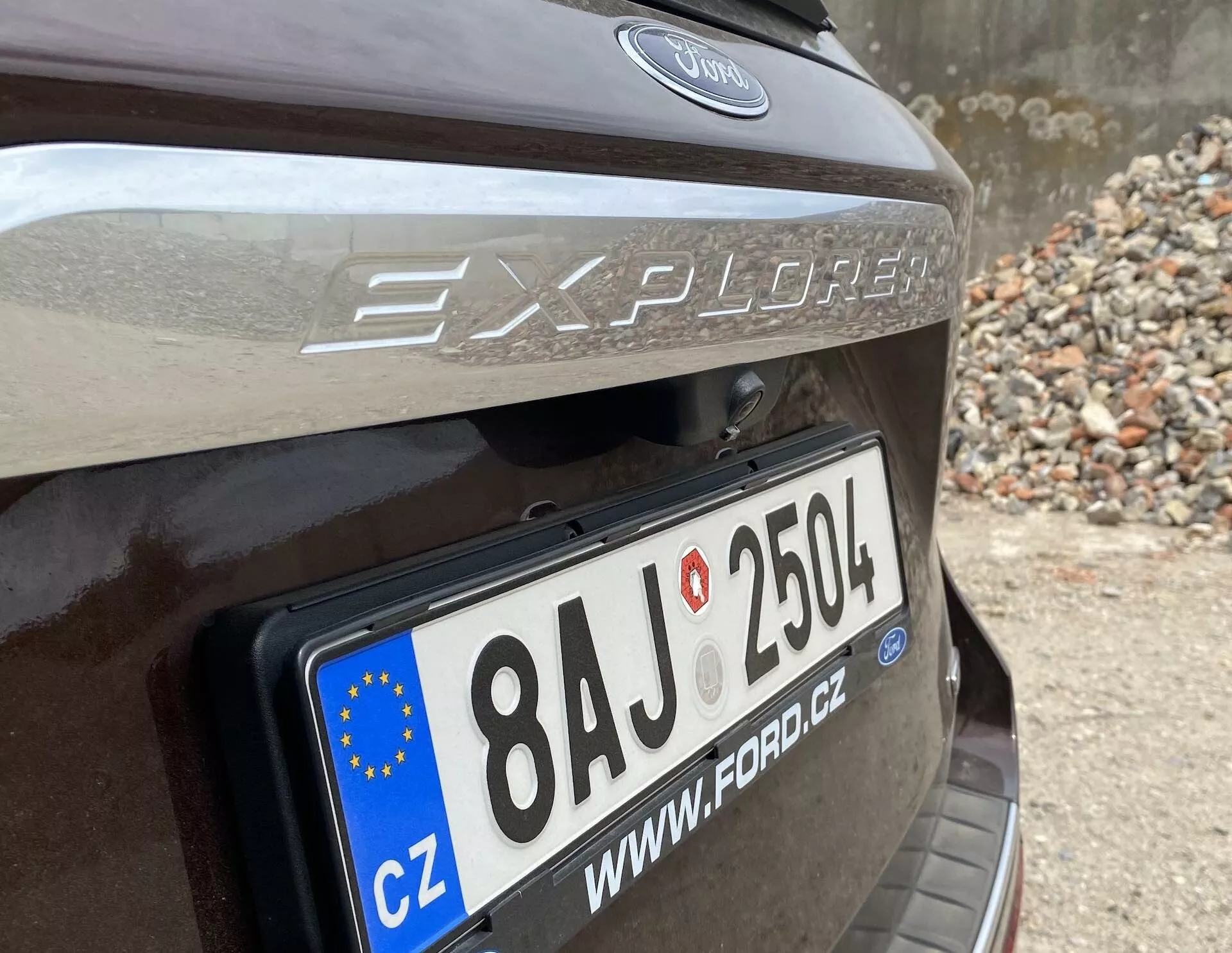 Närbild av bakdelen på en 2019 Ford Explorer, med fokus på bakluckans område med en framträdande EU-registreringsskylt som visar '8AJ 2504' och landskoden 'CZ'. Modellnamnet 'Explorer' och Ford-logotypen finns ovanför skylten, med en reflekterande remsa synlig ovanför registreringsskylten. Bilen verkar vara mörkbrun eller mörkgrå och parkerad i ett område med stenar eller spillror.